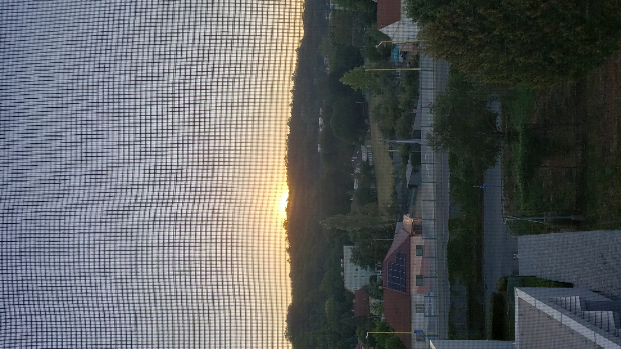 This image captures a sunset over a suburban landscape in Prešov, Slovakia. The foreground features a modern building with a white facade and a gray tiled walkway. Beyond this, a street is visible, bordered by a fence and lush green trees. Several houses with red roofs, some equipped with solar panels, are scattered throughout the midground. Further in the distance, a tree-covered hill rises, silhouetted against the warm orange and yellow hues of the setting sun. The sky above is a soft gradient of blue and gray. The view appears to be through a window screen, indicated by the faint grid pattern overlaying the entire image.