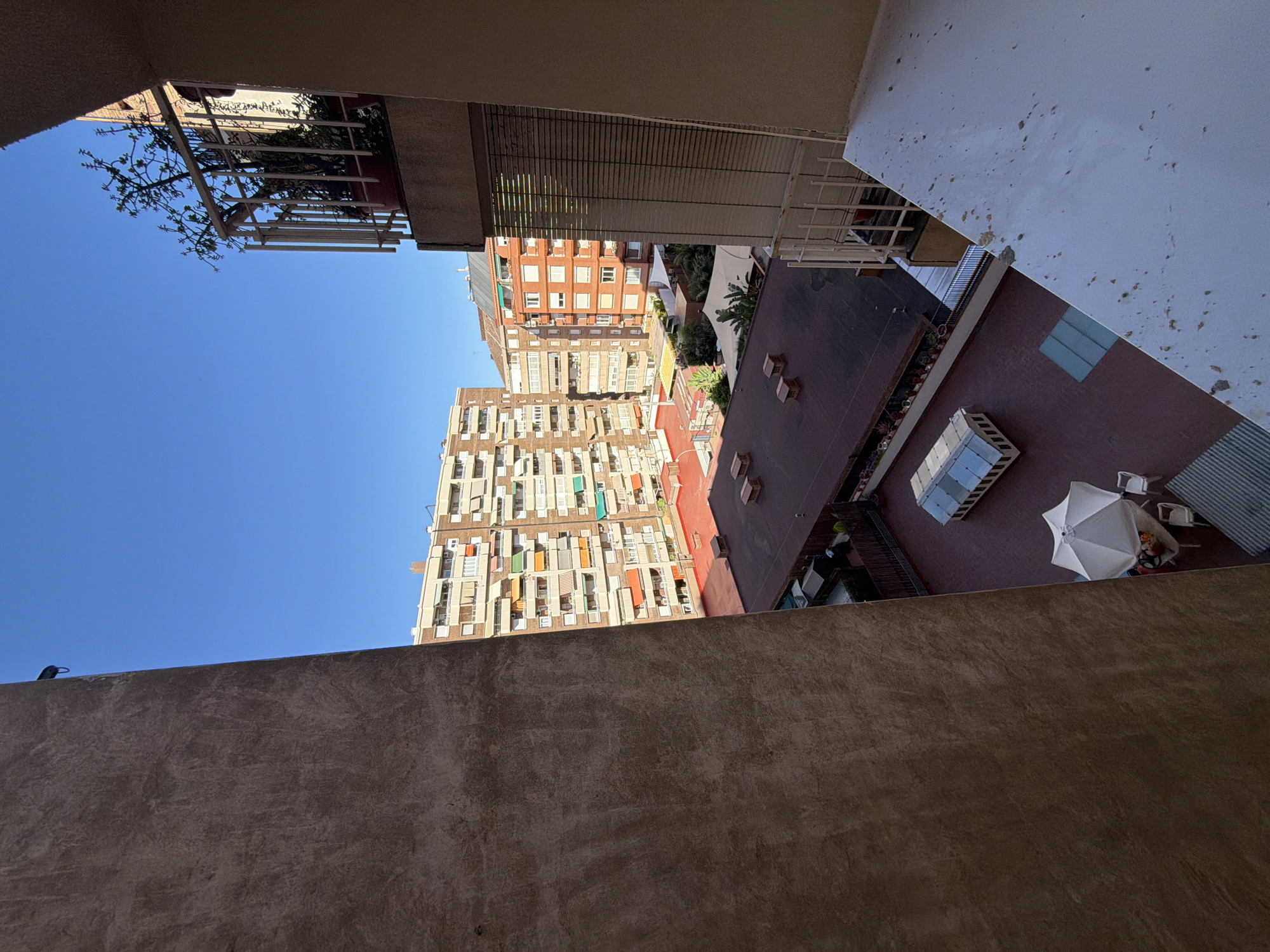 The image captures a view of an urban landscape, likely from a balcony or rooftop, in the Eixample neighborhood of Barcelona, Spain. The scene is dominated by residential buildings, characteristic of the area, with their distinct architectural styles and facades. The perspective looks down and across at the rooftops and balconies of surrounding structures, revealing a typical city vista.
In the foreground, a large, textured wall, possibly part of the building the photo is taken from, occupies the left side of the frame, casting a shadow. To the right, a balcony railing is visible, with a potted plant on it. Beyond this, a series of multi-story apartment buildings are seen. These buildings are characterized by rows of windows, many adorned with awnings in various colors, suggesting a sunny climate. The buildings appear to be constructed with brick and have balconies or terraces.
The lower portion of the image reveals rooftops with a reddish-brown surface, punctuated by ventilation shafts or small architectural elements. A white umbrella and chairs are set up on one of these rooftop areas, suggesting a small outdoor seating space. A glass-covered structure, perhaps a skylight or a small greenhouse, is also visible on one of the rooftops. The sky is a clear, bright blue, indicating a sunny day. The overall impression is one of a densely populated urban environment with residential living taking precedence.