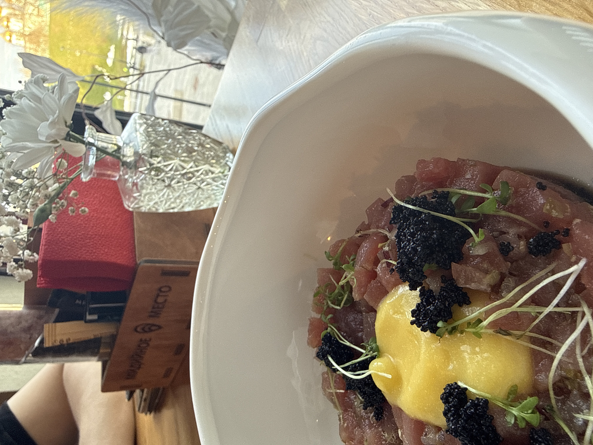 The image captures a close-up, slightly overhead view of a dining setting, likely in a restaurant or cafe in Dmitrov, Russia. The main subject is a white, irregularly shaped bowl holding a dish, prominently positioned on the right side of the frame. The dish consists of finely diced red meat, resembling tuna tartare, generously garnished with dark black caviar, a vibrant yellow, viscous sauce or egg yolk in the center, and delicate green microgreens or sprouts.
To the left, a floral arrangement adds to the decor, featuring several white daisy-like flowers and sprigs of white baby's breath, arranged in a textured, clear glass vase. Behind the vase, a red, textured object, possibly a folded napkin or book cover, is visible. Further left and below, a light-colored wooden stand or menu holder displays visible text. The words "КОЗЫРНОЕ МЕСТО" and "МЕСТО" are clearly discernible, translating to "Kozyrnoye Mesto" (meaning "Trump Place" or "Ace Place") and "Place," respectively, suggesting the name of the establishment or a phrase associated with it. A person's bare leg or thigh is partially visible at the bottom left, suggesting they are seated at the table. The background features a bright, blurred view through a window, indicating a daytime setting with yellowish-green light. The table surface is a light, possibly wooden, material. The overall scene suggests a pleasant dining experience.