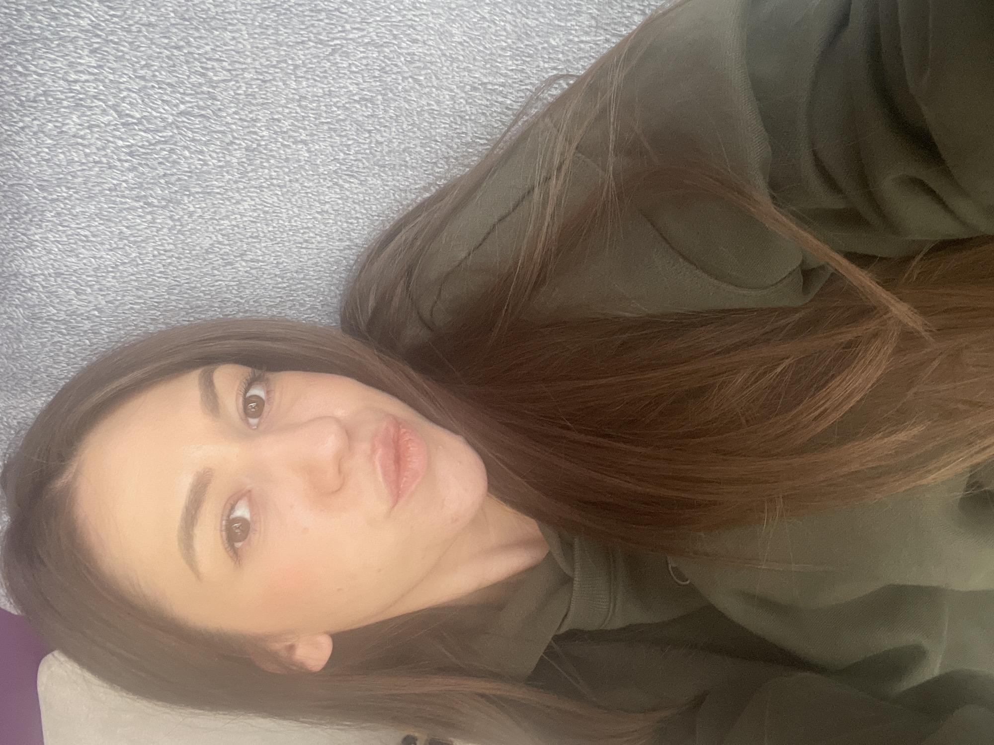 A young adult female with long brown hair is lying down, looking at the camera. She is wearing a green hoodie. The background is a grey textured surface. It is impossible to determine if this image was taken in Sofia, Bulgaria, based on the visual information provided.
