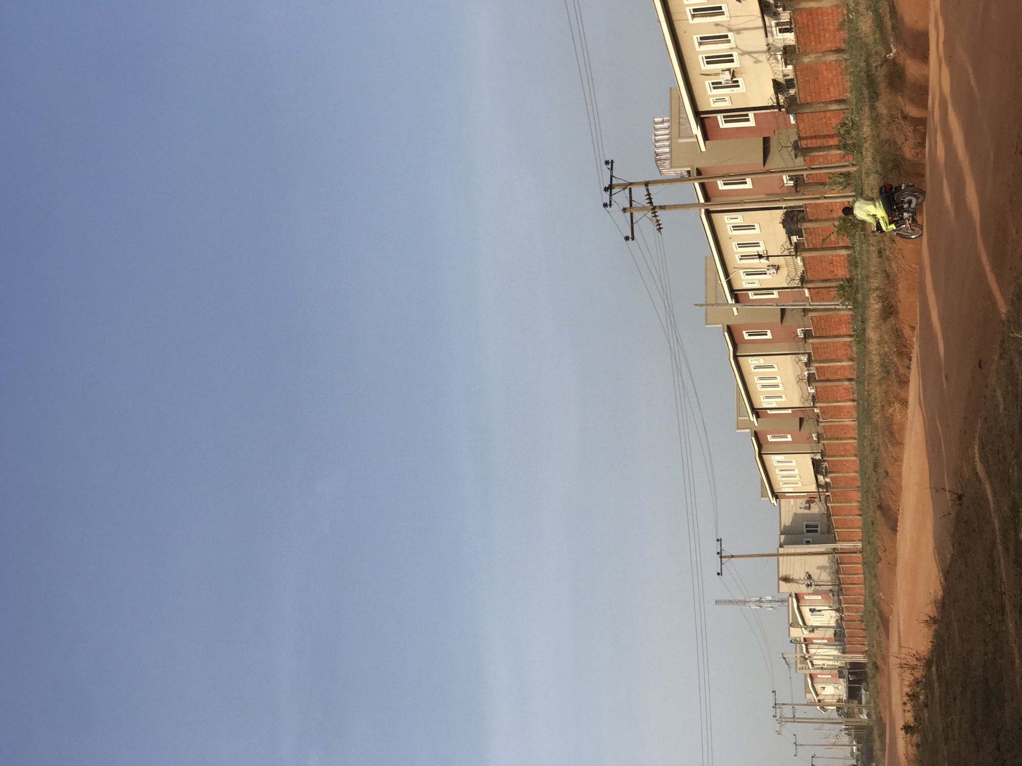 An outdoor scene captures a row of houses lining a reddish-brown dirt road under a clear, bright blue sky, characteristic of a daytime setting, likely late afternoon or early morning, given the soft lighting. The location is contextualized as Abuja, Nigeria.
On the unpaved dirt road, a person wearing a light green or yellow top is seated on a dark motorcycle or moped, facing away from the camera and positioned towards the right side of the road.
The houses are multi-story structures, primarily light beige or off-white, featuring dark roofs and distinct brick-red bases or lower wall sections. Many windows are visible, framed in white, and some buildings show external air conditioning units. Between properties, brick walls topped with barbed wire can be seen, indicating boundary divisions and security. A series of wooden electricity poles, laden with multiple overhead wires, run parallel to the road and houses, providing power infrastructure. Further down the street, a tall communications antenna or mast rises above the buildings. The terrain alongside the road and between houses consists of dry, sparse grass and scrubland, suggesting a dusty, possibly suburban or developing area. There is no visible text in the image.