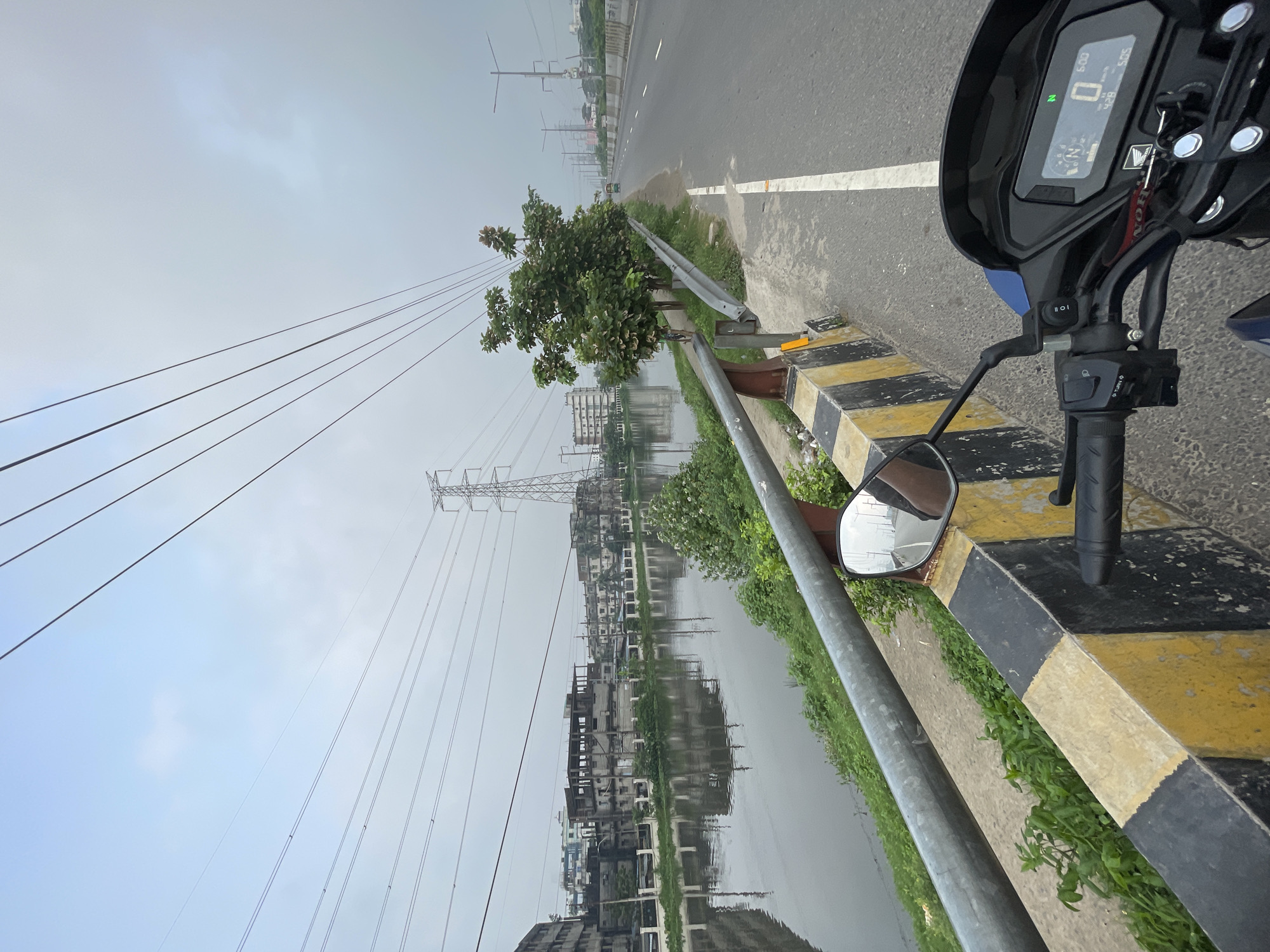 The image captures a scene in Dhaka, Bangladesh, specifically from the perspective of a person on a motorcycle riding along a road bordering a body of water. The motorcycle is partially visible in the foreground, including the dashboard indicating a speed of 0 km/h and the rearview mirror.
A gray metal guardrail runs along the road's edge, marked with alternating black and yellow stripes. Beyond the guardrail, the water reflects the overcast sky and the silhouettes of buildings, some of which appear to be under construction.
Overhead, high-tension power lines run parallel to the road, supported by metal transmission towers. A small tree is planted near the guardrail. The road is paved and marked with a solid white line. The sky is overcast, indicating a cloudy day. The surrounding environment features vegetation growing between the guardrail and the water's edge. The scene suggests an urban or suburban area undergoing development, with a mix of infrastructure and natural elements.
