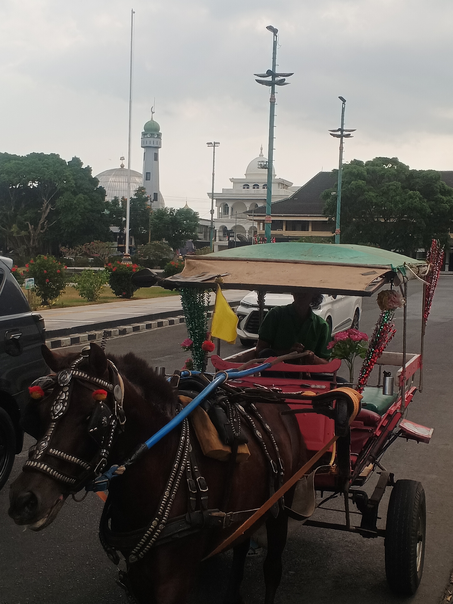 In Sokanegara, Indonesia, a horse-drawn carriage, known locally as a "andong," is parked on a street. The horse, a brown mare, is adorned with ornate silver and red embellishments on its bridle and harness. It is attached to a red carriage with a green canvas roof. A man wearing a green shirt is seated in the carriage, holding the reins. In the background, a white mosque with a green dome and a tall minaret stands prominently. The sky is overcast. Several trees and a white car are also visible in the scene.