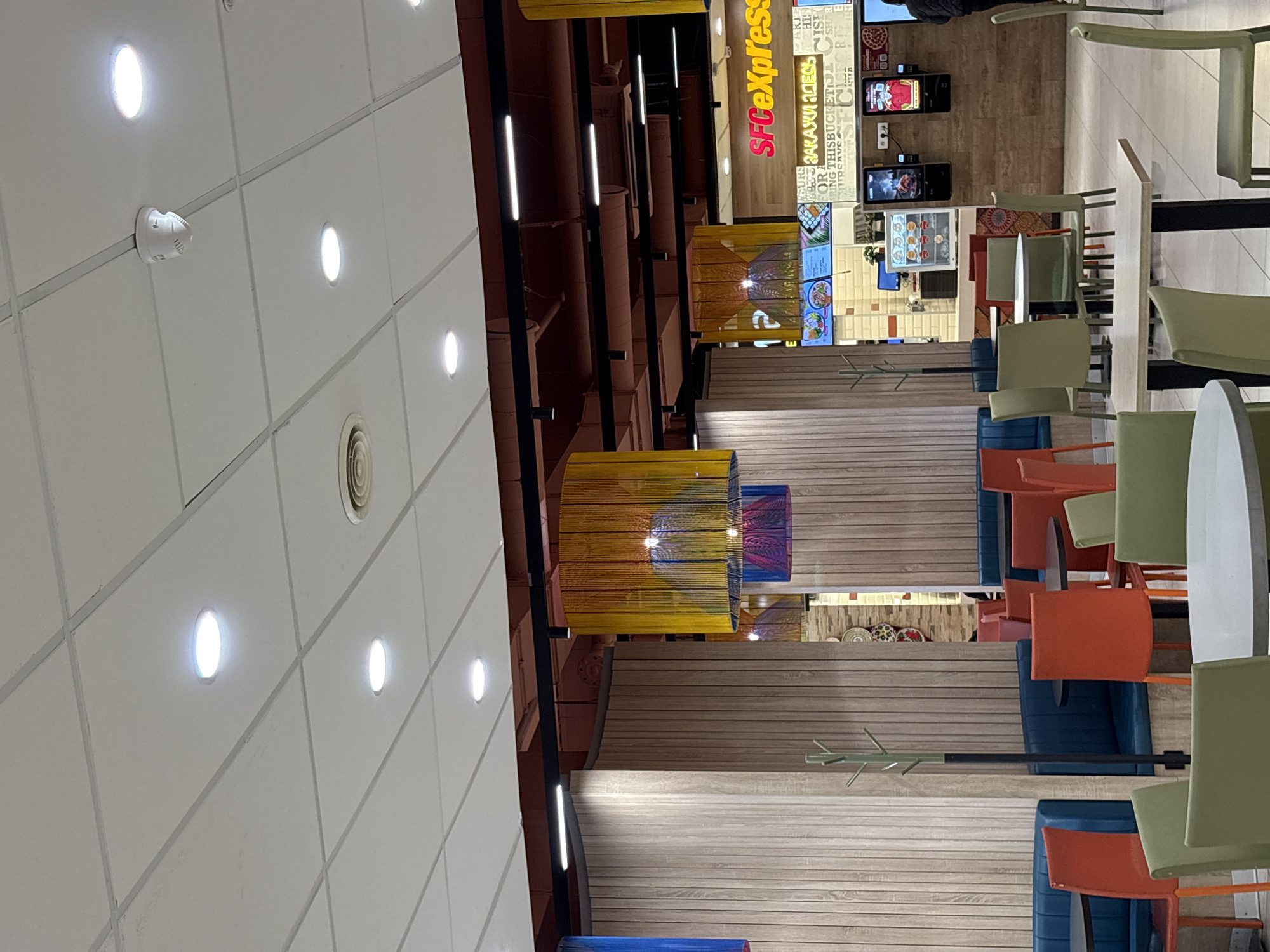 The image shows the interior of a food court or dining area in an airport or similar public space. Overhead are fluorescent lights set in a grid on a white ceiling, along with ceiling-mounted fixtures such as a smoke detector and air vent.
To the right of the ceiling, a series of dark horizontal bars run along the space, which form part of the structural or decorative design. Hanging from the ceiling structure are a series of yellow and blue woven light fixtures.
The lower half of the image shows seating arrangements. There are a variety of chairs in shades of green, orange, and grey, arranged around tables. There are also rows of built-in seating upholstered in blue. A coat rack stands against a wall clad with horizontal wooden planks.
Toward the upper right corner, the signage for "SFCExpress" is visible above a food vendor's display. Menu boards and digital displays are visible in the vendor area. In the extreme upper right, people stand in line.