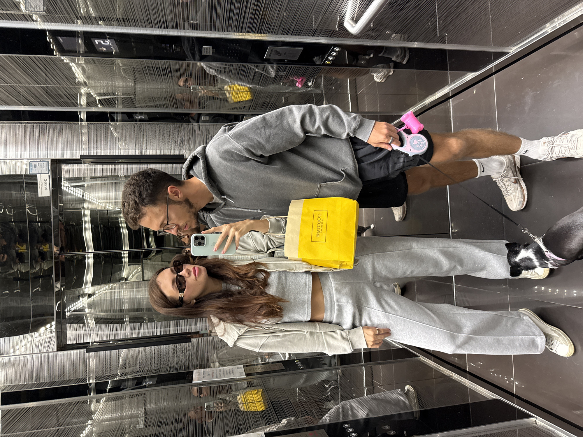 A couple in casual clothing is taking a selfie in an elevator in Tel Aviv, Israel, accompanied by a dog. The elevator's interior is notable for its reflective surfaces and decorative vertical strands. The woman wears sunglasses and grey sweatpants with a matching top and holds a shopping bag with the name L'OCCITANE en Provence visible. The man, wearing glasses and a grey hoodie, is holding a dog waste bag dispenser. The dog is on a leash, and the elevator's panel and button details are visible in the reflection.