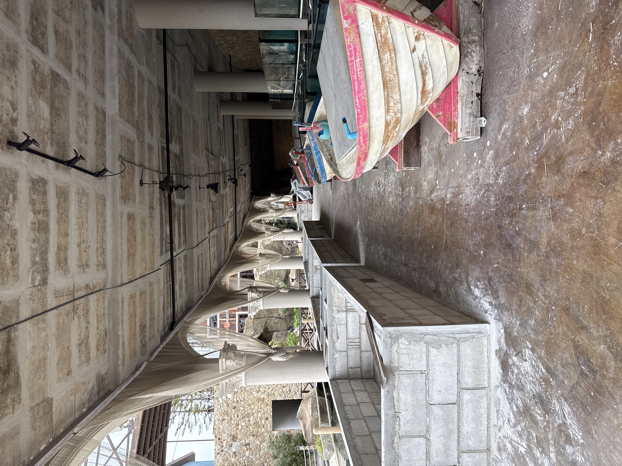 The image depicts an outdoor area in Pattaya City, Thailand, likely a construction or renovation site adjacent to an aquarium or aquatic facility. In the foreground, several unfinished concrete block enclosures line the left side of the image, some containing wooden planks. To the right, a row of small, weathered boats rests on wooden stands. The boats appear to be fiberglass or wooden, with chipped paint and visible wear. The foremost boat is white with prominent pink trim along its upper edge, and a blue pipe extends from its interior. Behind it, other boats are visible, with at least one featuring red and blue trim. The ground is a worn concrete or asphalt surface, showing signs of dust and debris. Overhead, a textured concrete ceiling with exposed beams is supported by large cylindrical columns. Several industrial-style lights are suspended from the ceiling. Draped fabric or netting hangs from arched structures along the left side, adding to the unfinished aesthetic. In the background, a stone wall and some greenery are visible, along with large glass tanks, suggesting the presence of an aquarium. The overall scene is one of ongoing construction or a state of disuse, with a focus on the boats and the structural elements of the environment.