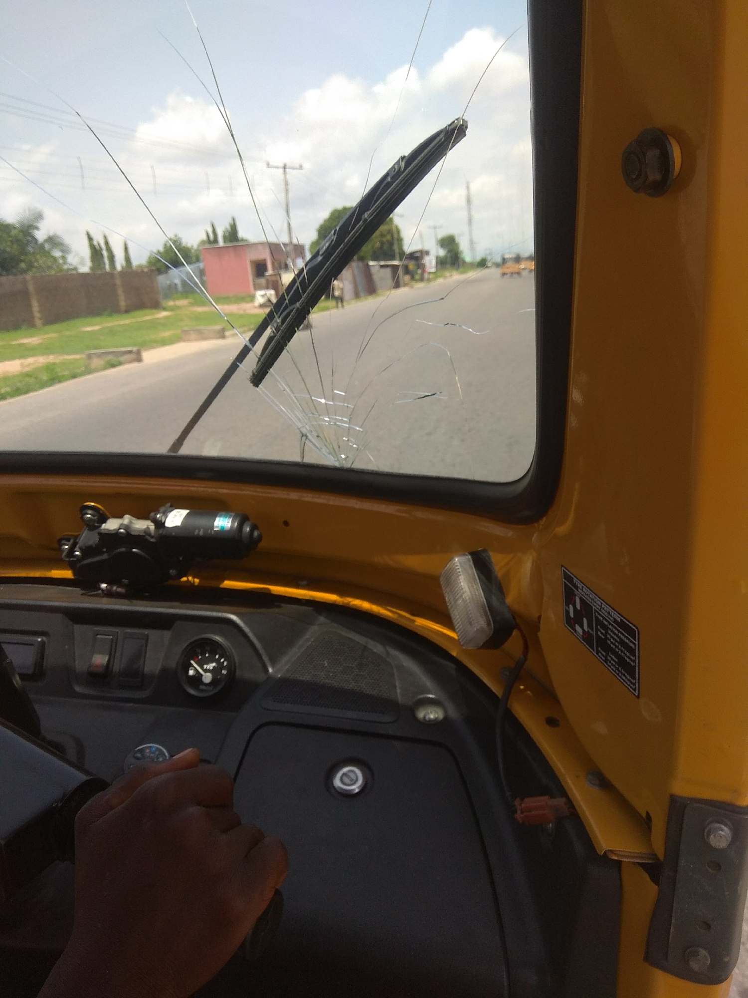 The image depicts the interior of a yellow auto-rickshaw, viewed from the driver's perspective. The windshield is visibly cracked, with several long, spiderweb-like fractures extending across it. A windshield wiper is attached to the top of the windshield. In the foreground, the driver's dark-skinned hand is seen gripping the steering wheel. The dashboard features several controls, including switches and a circular gauge. A windshield wiper motor is mounted on the yellow casing above the dashboard.
The background, visible through the cracked windshield, shows a paved road with sparse vegetation and buildings lining the sides. Power lines and utility poles are also visible. The sky is partly cloudy. A few other vehicles, including what appears to be another auto-rickshaw, can be seen in the distance on the road. The overall scene suggests an outdoor environment in a city.
Based on the visual cues of the auto-rickshaw, the style of the buildings, and the type of vegetation, this scene is consistent with an urban or semi-urban environment in Nigeria, specifically Jalingo. The cracked windshield may indicate a recent incident, such as a collision or damage from debris.