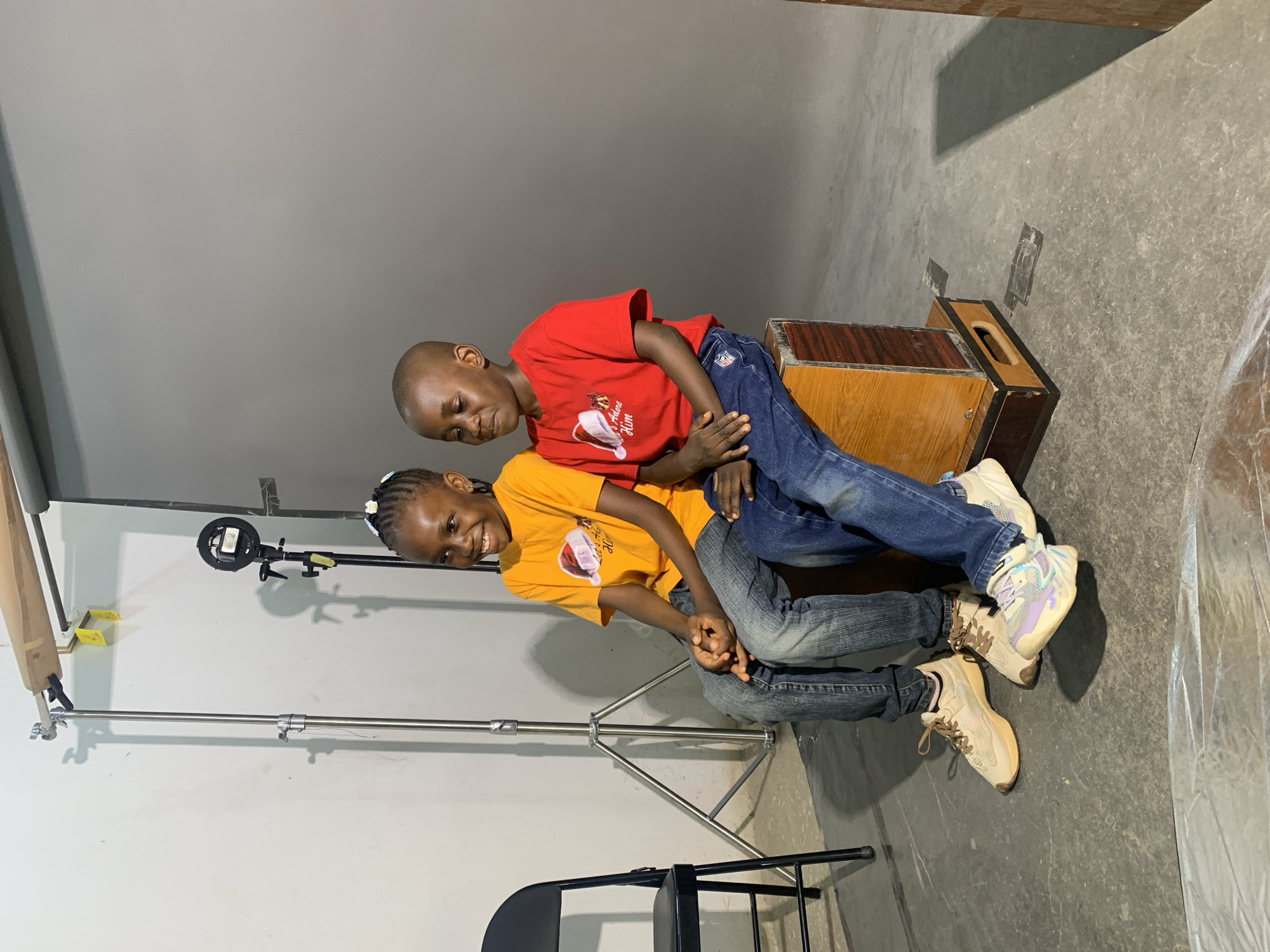 In a studio setting in Gura Top, Nigeria, a young boy in a red t-shirt and jeans sits on a wooden box, leaning on a smiling young girl wearing an orange t-shirt and jeans. The girl sits on a black chair in front of a photography backdrop stand. The studio has a gray floor and walls, with photography equipment visible.
