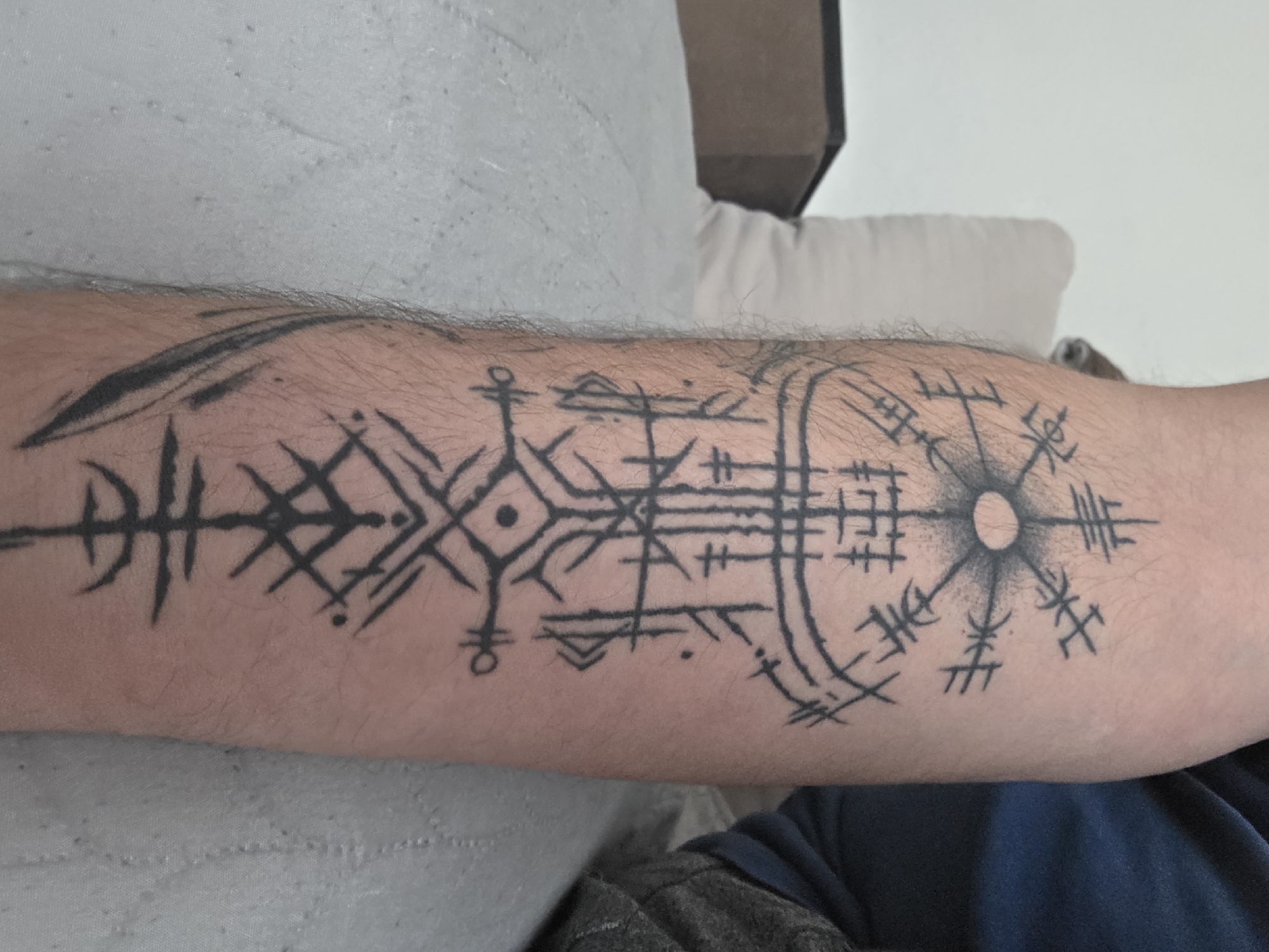 A forearm is tattooed with black ink in a complex design incorporating runes, circles, and geometric shapes. The skin on the forearm is fair and covered in fine hairs. The forearm rests on a grey textured surface, which appears to be fabric. Behind the forearm is a white cushion and a dark rectangular shape.
The location in the city of Blagoevgrad, Bulgaria is not ascertainable from this image.