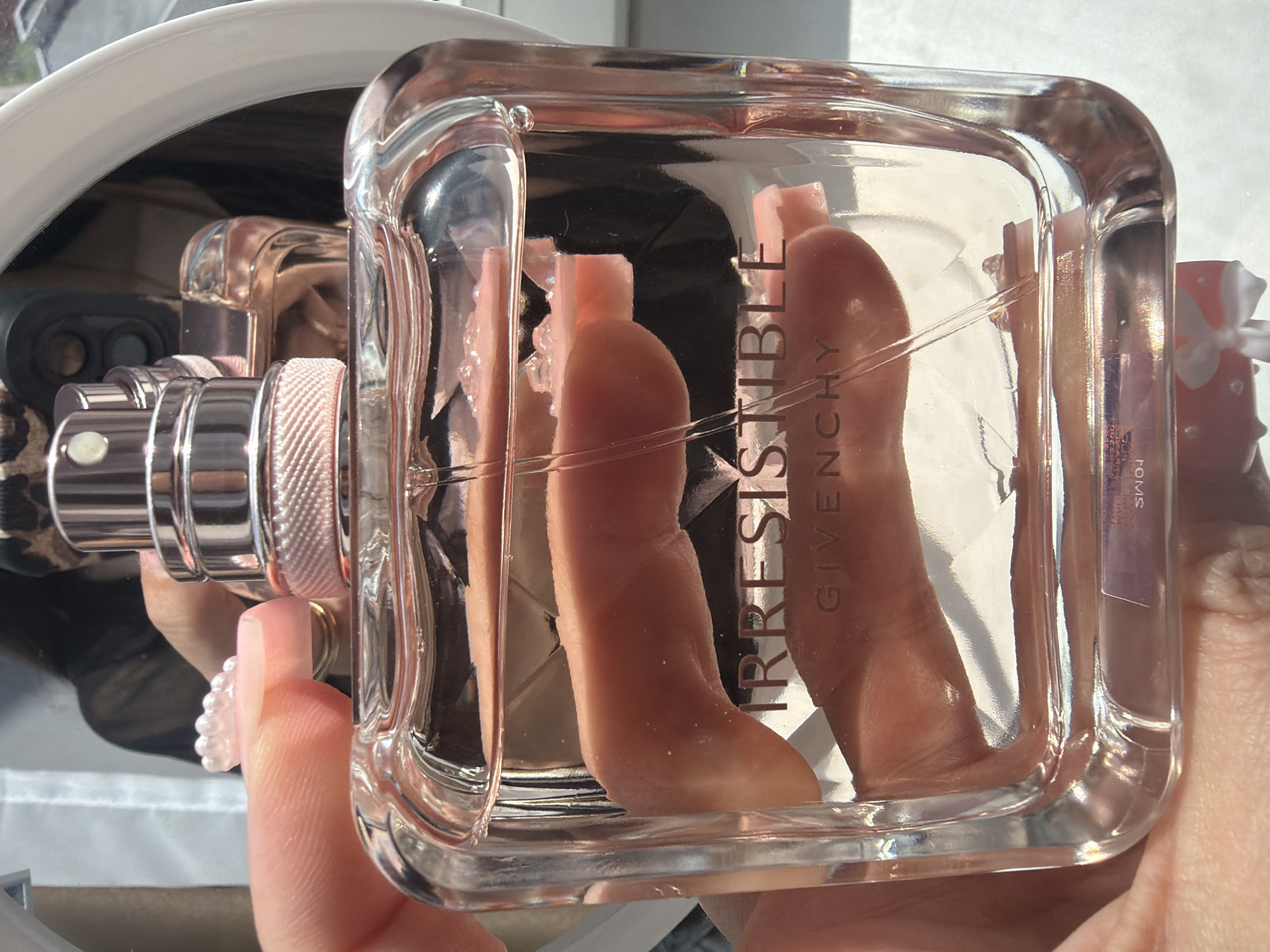 A close-up, brightly lit image features a right hand holding a clear glass perfume bottle containing a pink-tinted liquid, approximately three-quarters full. The bottle's front is embossed with the word "IRRESISTIBLE" in prominent dark, reflective capital letters, with "GIVENCHY" inscribed below it in a smaller font. The top of the bottle displays a metallic rose gold and silver sprayer mechanism, detailed with a textured, light pink band below the cap and a clear plastic dip tube extending into the fragrance. The hand exhibits manicured nails with a clear, iridescent beaded embellishment on the index finger. A thin gold-colored ring is visible on another finger. In the highly reflective background, partially seen within a white-framed mirror, is the reflection of a person wearing dark, possibly patterned clothing, holding a dark electronic device, likely the camera used for the photograph. Strong, directional lighting, possibly natural sunlight, creates numerous highlights and reflections across the bottle's surface. The location is in the city of Novosaratovka, Russia.