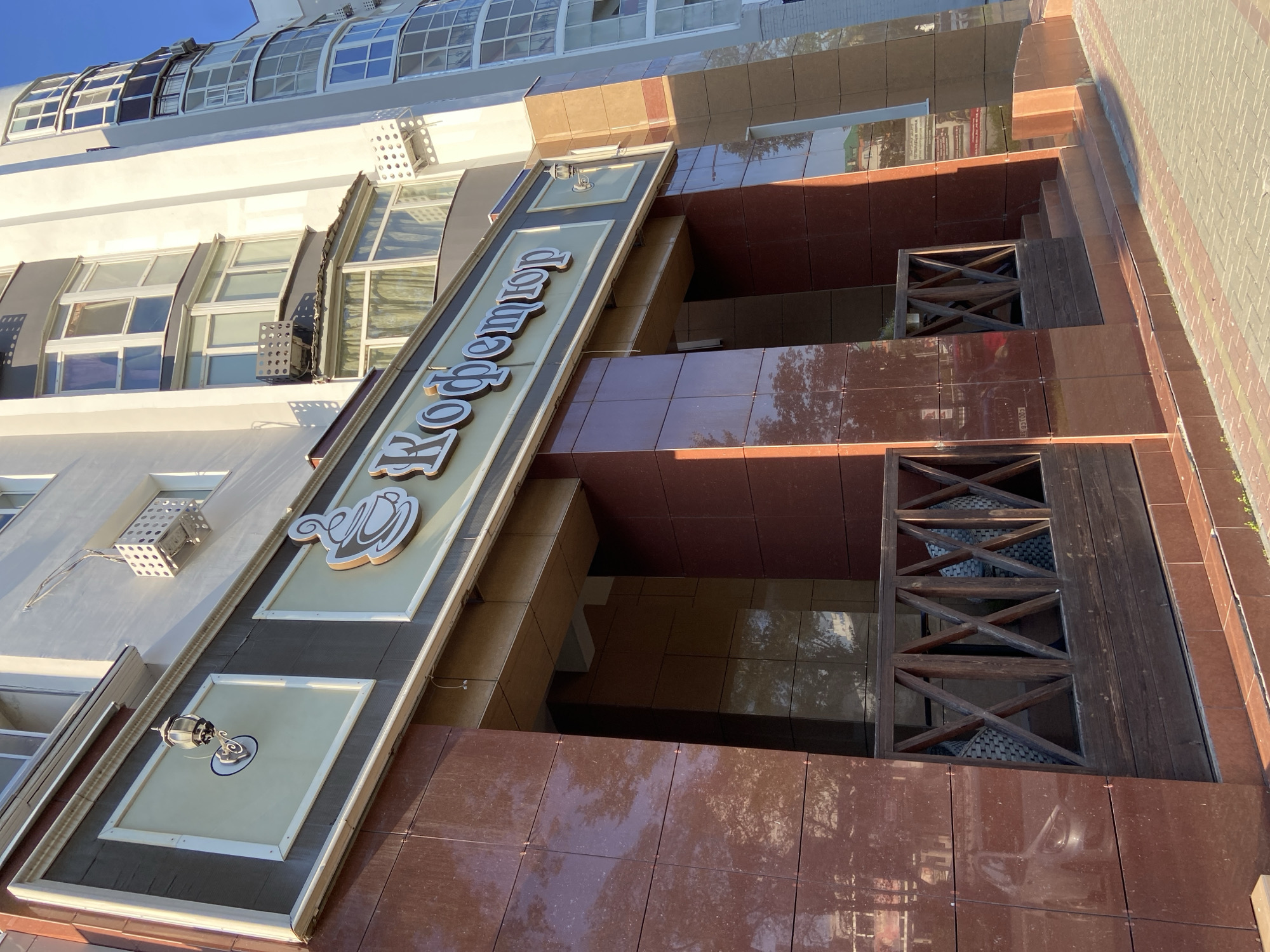 The image shows the exterior of a cafe named "Кофетор" (Kofetor) located in Blagoveshchensk, Russia. The cafe has a modern facade with large, reflective brown marble tiles on the lower section and a lighter-colored upper section. Above the entrance, there is a prominent sign with the cafe's name and a stylized coffee cup logo. The cafe has a wooden fence enclosing a seating area on the ground floor. The building itself appears to be part of a larger residential complex, with visible windows and balconies on higher floors. The scene is set outdoors on a sunny day, with shadows indicating the time of day. The foreground shows a paved sidewalk. There are no people visible in the image.