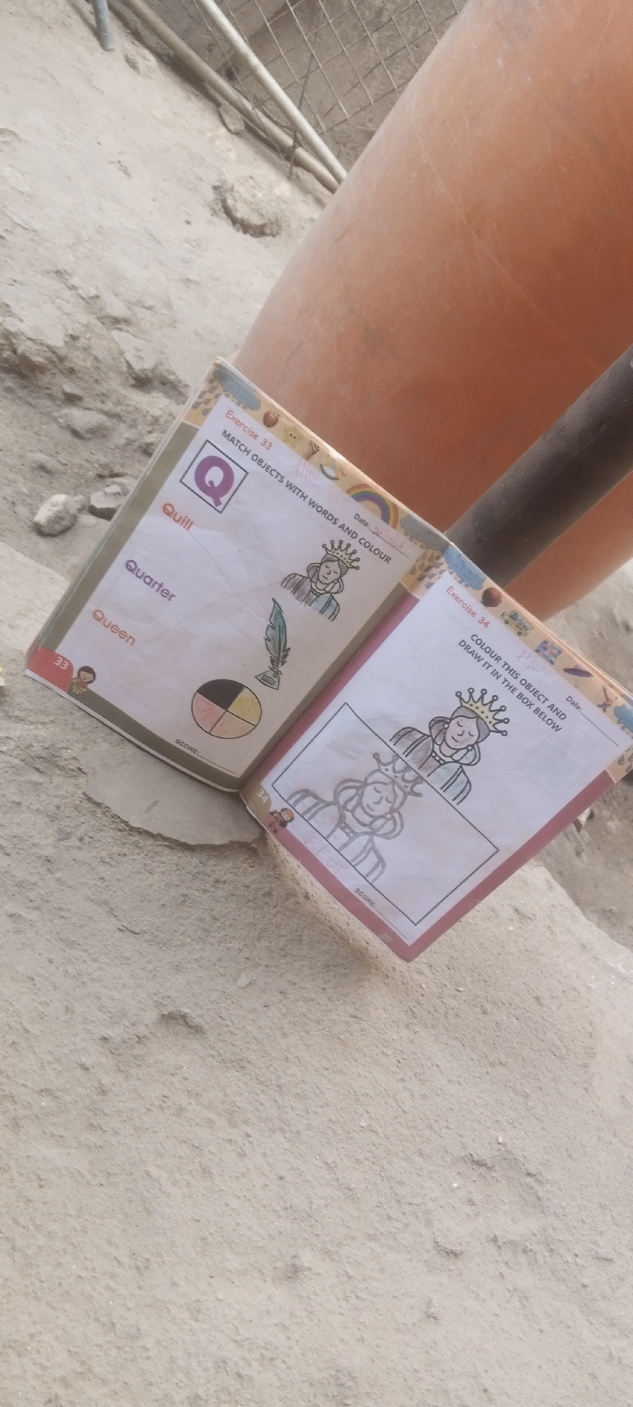 The image shows an open children's activity book, with pages titled "Exercise 33" and "Exercise 34," lying on a dirt surface. The pages are related to matching objects with words and coloring, featuring illustrations of a queen, a quill, and a quarter. The location is implied to be Sabon Tasha, Nigeria. The environment includes a dirt surface and a blurred view of an orange cylindrical object.