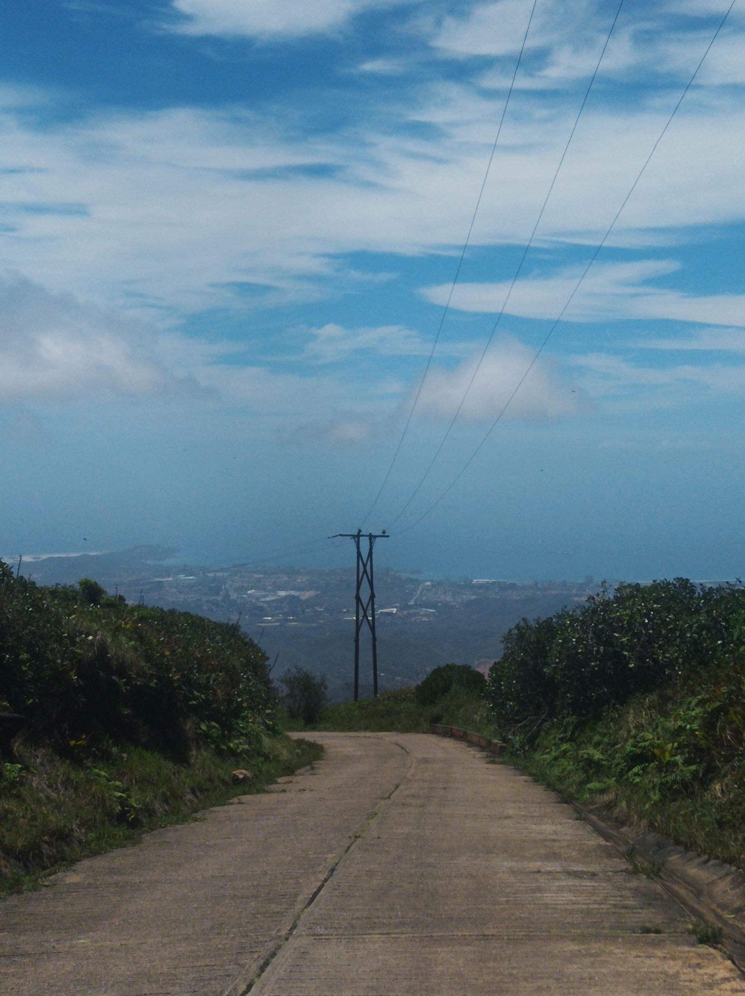 A medium-angle shot depicts a concrete road leading toward a distant cityscape. The road, cracked in places, is framed by lush green vegetation on both sides. Overhead, power lines stretch from the top of the frame toward a utility pole in the middle distance. Beyond the pole, a hazy view of Las Piedras, Venezuela, and the sea stretches to the horizon under a sky with scattered clouds.