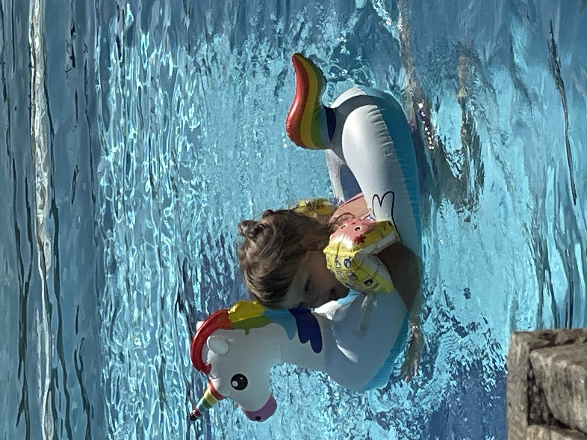 A young child with brown hair pulled back is floating in a bright blue swimming pool on an inflatable unicorn. The child is wearing yellow arm floaties with a pink design and appears to be smiling and enjoying themselves. The unicorn float is white with a rainbow mane and tail, and a rainbow horn. The water is clear and reflects the sunlight, creating ripples and shimmering effects. In the foreground, a portion of a stone border is visible. The scene depicts a recreational activity in a swimming pool.