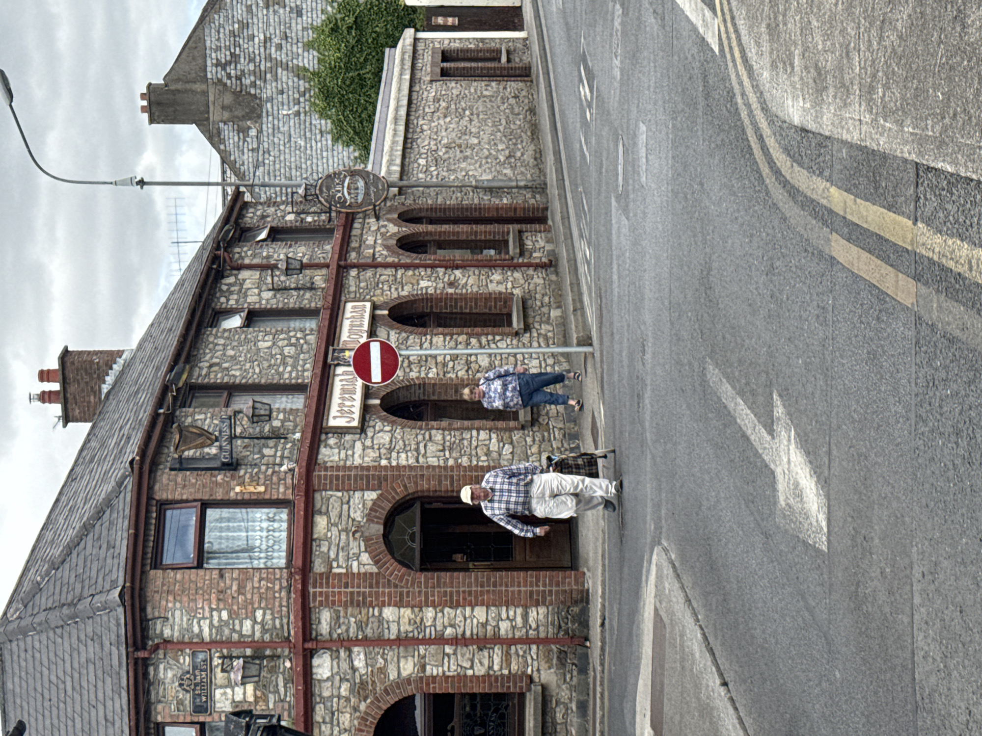 The image depicts a street scene in Clonmel, Ireland, focusing on the exterior of a traditional stone building identified by signage as "Jeremiah Moynihan." The building has a stone facade with red brick accents around arched windows and doorways. A Guinness harp logo is visible on the upper left side of the building, indicating it is likely a pub or establishment serving the beverage.
In the foreground, a man in a checkered shirt and light-colored pants walks across the asphalt road, pulling a wheeled shopping cart. Further down the street, a woman in a floral patterned top and blue jeans walks in the same direction. A circular red and white traffic sign, indicating "no entry" or a similar prohibition, stands on a pole near the building. The street is paved with asphalt and marked with white directional arrows. The environment is overcast, suggesting a cloudy day. Buildings with stone and brick construction line the street, with some greenery visible in the background. The overall impression is a street in a historic Irish town.