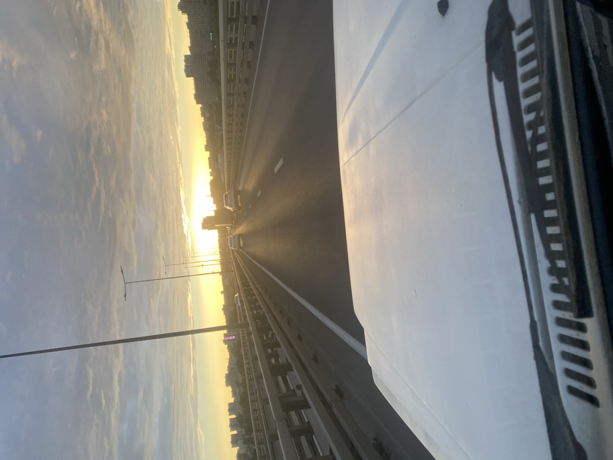 A car is driving on a bridge over a river during sunset in Nizhny Novgorod, Russia. The sun is setting on the horizon, casting a warm glow over the city. There are several other cars on the bridge, and buildings can be seen in the distance. The sky is cloudy, with streaks of orange and pink. The car's hood and windshield wipers are visible in the foreground.