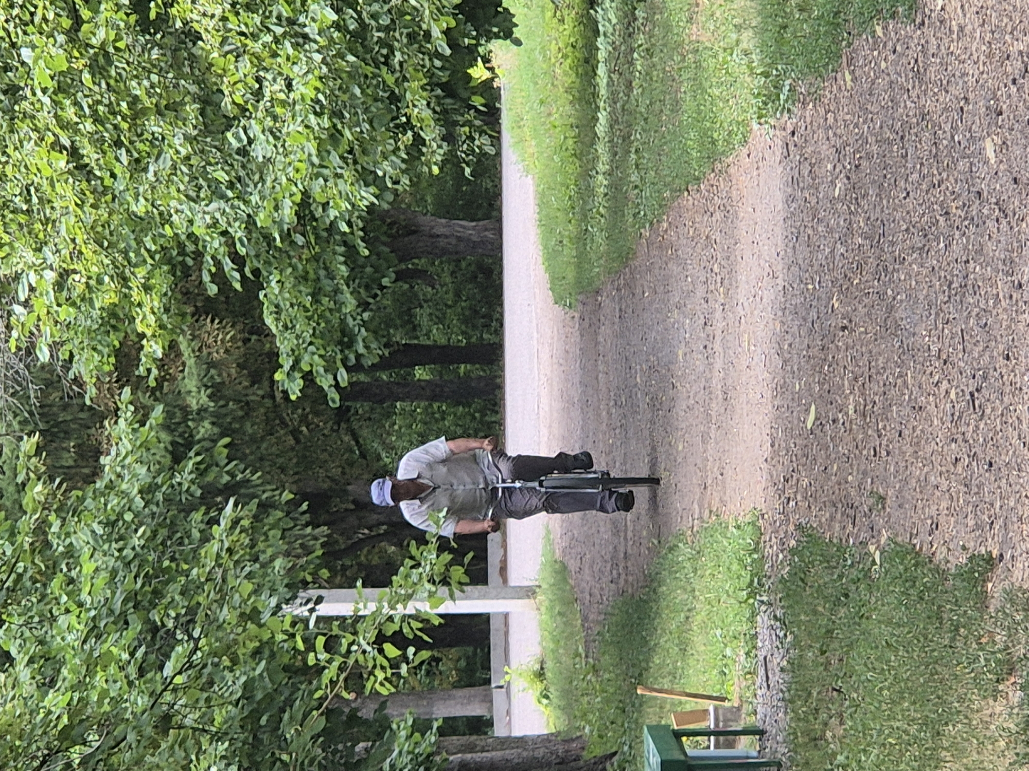 An outdoor, eye-level, full-shot photograph taken in Sozonivka, Ukraine, shows a man riding a bicycle on a gravel path that curves to the right. The man, wearing a light-colored collared shirt, dark pants, and a blue cap, is positioned centrally on the path, facing the camera. He is riding a dark-colored bicycle, and his hands are on the handlebars. The path is bordered on the left by grass and trees with dense green foliage. On the right, the path is also bordered by grass and more trees. A light-colored pole and some green benches are visible on the left side of the path in the foreground. The background consists of more trees and foliage, suggesting a park or a wooded area. The lighting is even, indicating an overcast day.