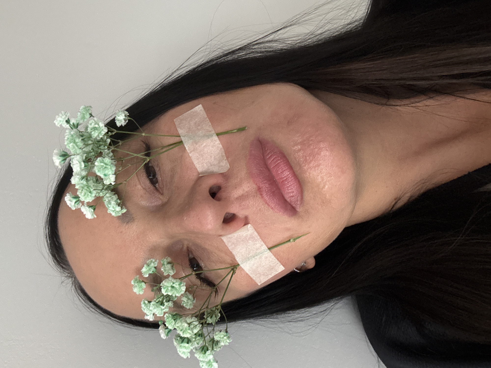 This media file presents an artistic close-up portrait of a person, likely a woman, in Chaikovsky, Russia. The individual's head is tilted to the left, with long, dark hair framing the face against a plain, light grey background, suggesting an indoor setting.
Small clusters of light green and white baby's breath flowers are strategically placed on the face, held in position by strips of light-colored adhesive tape. One arrangement of flowers is visible near the left eye and temple, secured by tape across the forehead. Another cluster is located on the right cheek, fastened with tape spanning the cheekbone. The person's lips are made up with a pinkish-mauve lipstick, and their eyes are open, gazing with a neutral expression. A subtle earring is visible on the right ear. The composition and placement of the flowers and tape suggest a deliberate, artistic pose. No text is visible in the image.
