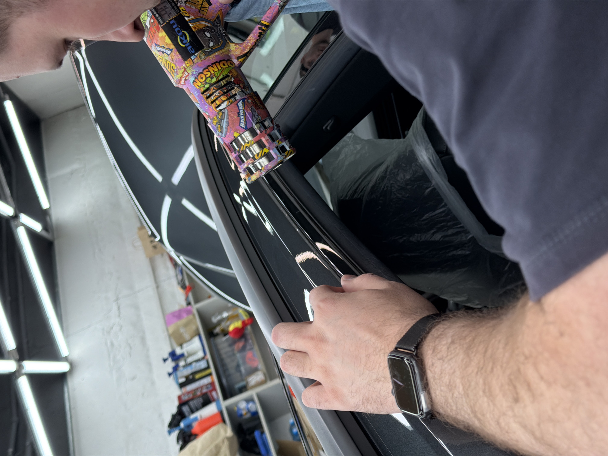 A man is working on a car inside a garage. The man wears a gray T-shirt and a black smartwatch on his left wrist. He is holding a heat gun covered in cartoon stickers and using it on the car. The car appears to be gray and has a glossy finish. The garage has a white brick wall with fluorescent lights, and a shelf filled with various tools and supplies is visible in the background. The location could potentially be in Krasnoyarsk, Russia, but this is not verifiable from the image alone.
