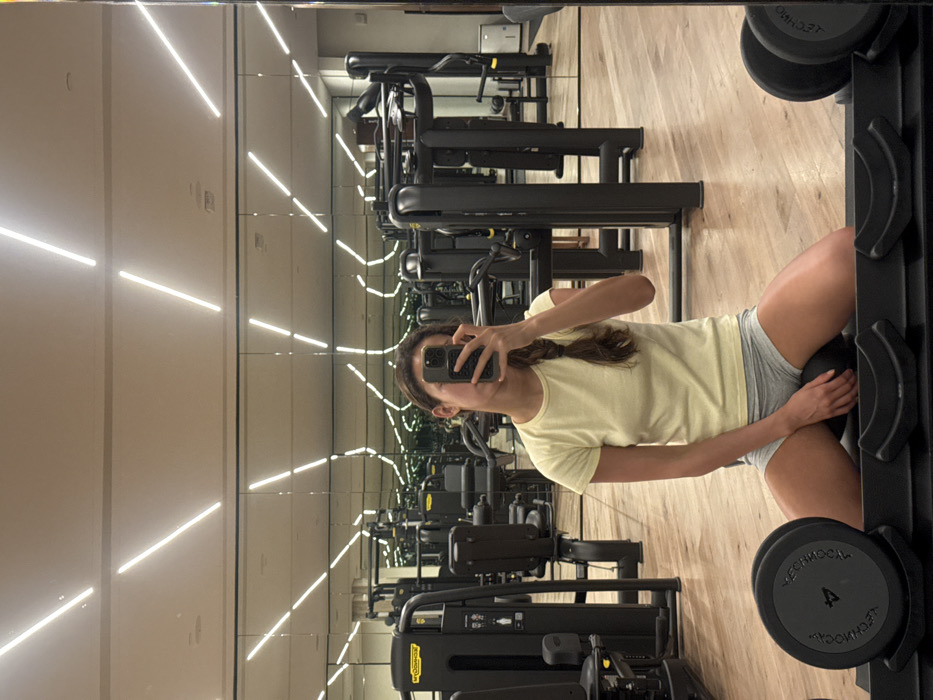 A young woman with braided hair is reflected in a gym mirror while taking a selfie with her phone. She wears a pale yellow t-shirt and grey athletic shorts. She is standing at a dumbbell rack holding a medicine ball between her thighs. Behind her, the gym is equipped with rows of weight machines and bright overhead lighting. The floor appears to be a light wood-tone.
