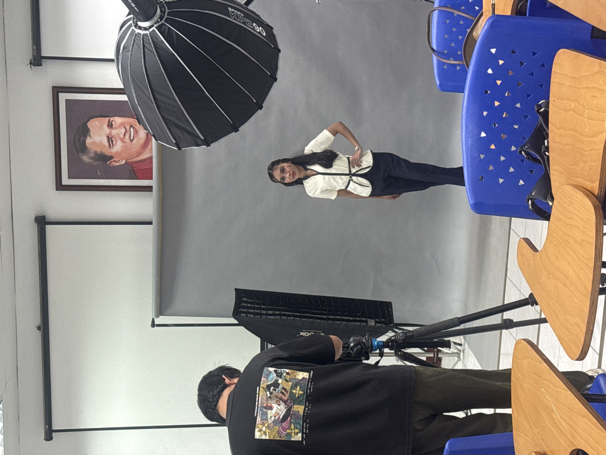 In a studio setting, possibly within an educational institution in Davao City, Philippines, a photo shoot is underway. A young woman with long, dark hair stands centrally against a gray backdrop, posing with her hand on her hip. She is wearing a white short-sleeved blouse with a dark trim and black pants. To her left, a photographer, viewed from behind, is focused on adjusting equipment. He wears a black t-shirt with a colorful graphic print on the back and dark trousers. In the foreground, two wooden desk surfaces with rounded edges are visible, accompanied by blue plastic chairs with triangular cutouts in their backs. A large softbox light stands to the left of the woman, and a parabolic umbrella light is positioned above and to her right, indicating a professional lighting setup. A framed portrait of a man with a contorted facial expression hangs on the wall behind the woman. The overall environment suggests a controlled studio space, possibly for formal portraits or a school-related event.