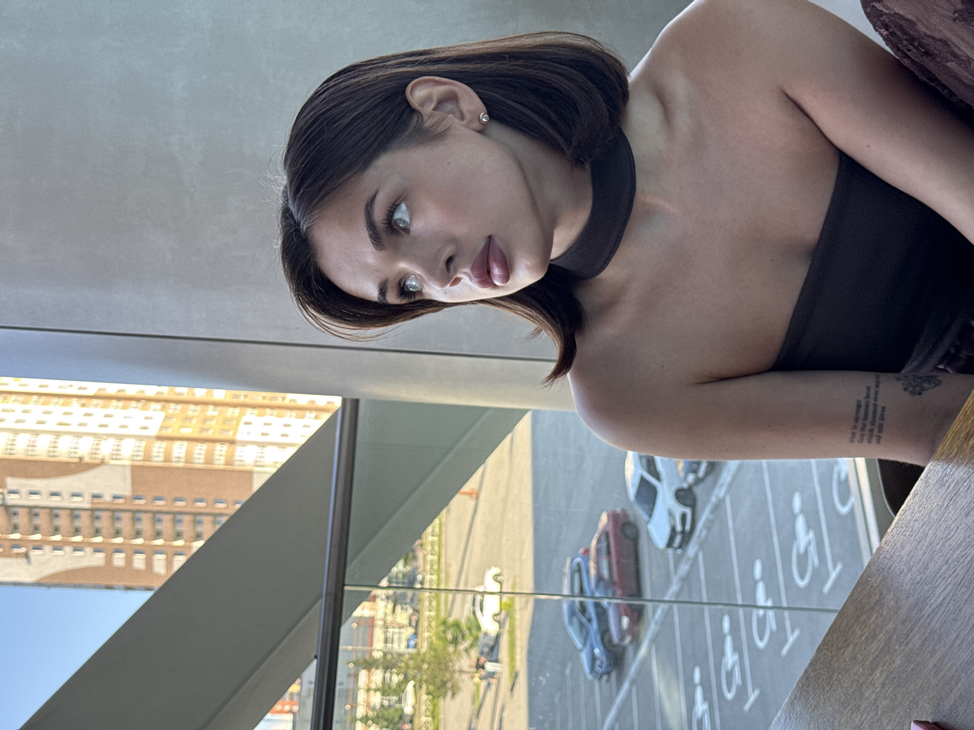 A young woman with shoulder-length brown hair and fair skin is pictured in a close-up shot, looking to her left. She is wearing a dark strapless top and a dark choker necklace. A delicate silver earring is visible in her right ear. Her left arm, resting on a wooden table, shows a tattoo with text and floral imagery. Through a large window behind her, a cityscape is visible, featuring a tall, modern apartment building with balconies and orange-tinted brickwork. The window overlooks a parking lot with several cars and designated parking spaces, including some marked for disabled individuals. The scene is bathed in natural light, suggesting it is daytime. The overall ambiance is one of calm observation. The architectural style and the tall residential building are consistent with urban environments in cities like Perm, Russia, though the exact location is not definitively identifiable from the image alone.