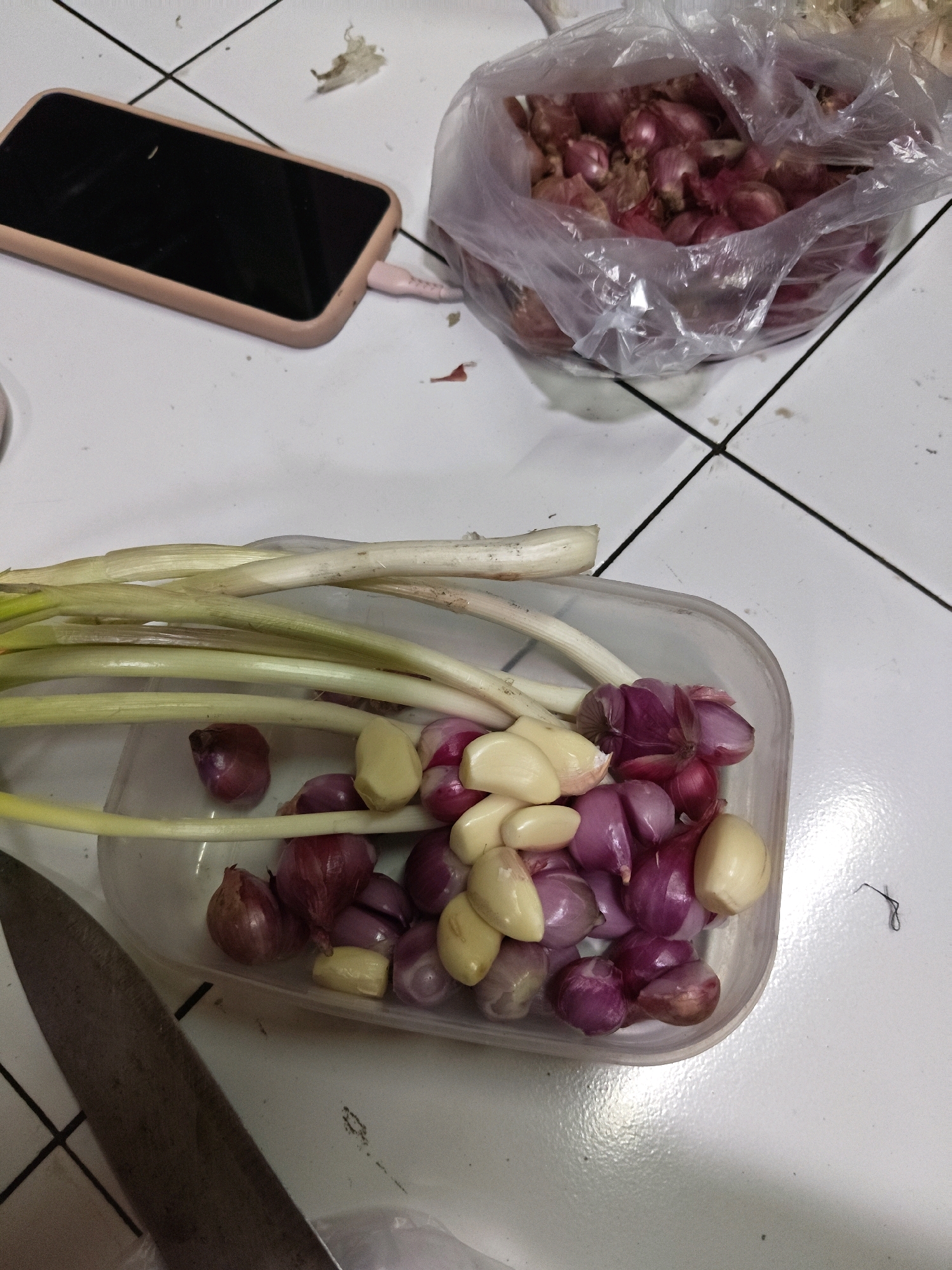 In Sumberwono, Indonesia, a close-up, overhead shot captures a collection of ingredients on a tiled surface. To the left, a smartphone with a pink case is plugged in. A plastic bag filled with red onions is in the upper right corner. In the foreground, a clear plastic container holds peeled garlic cloves and more red onions, alongside what appear to be scallions. A metal knife lies to the bottom left. The scene is dimly lit, suggesting an indoor environment.