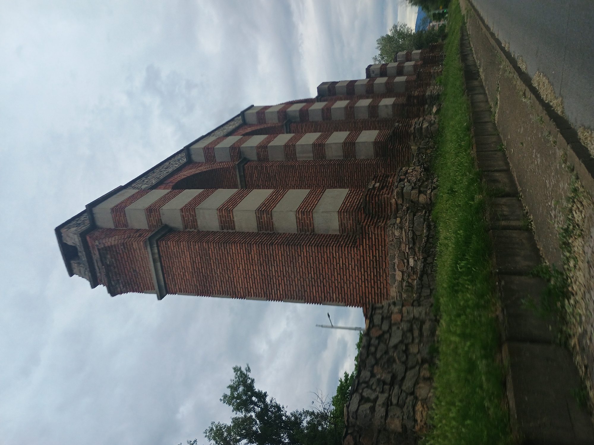 The image shows the Water Tower, located in Giurgiu, Romania. It is a tall, brick structure with alternating bands of red brick and light-colored stone or concrete. The tower features a distinct stepped design near the top, where it progressively narrows. A portion of the stone base of the tower is visible. The tower is bordered by grass and a sidewalk. A road is adjacent to the sidewalk. The sky is overcast.