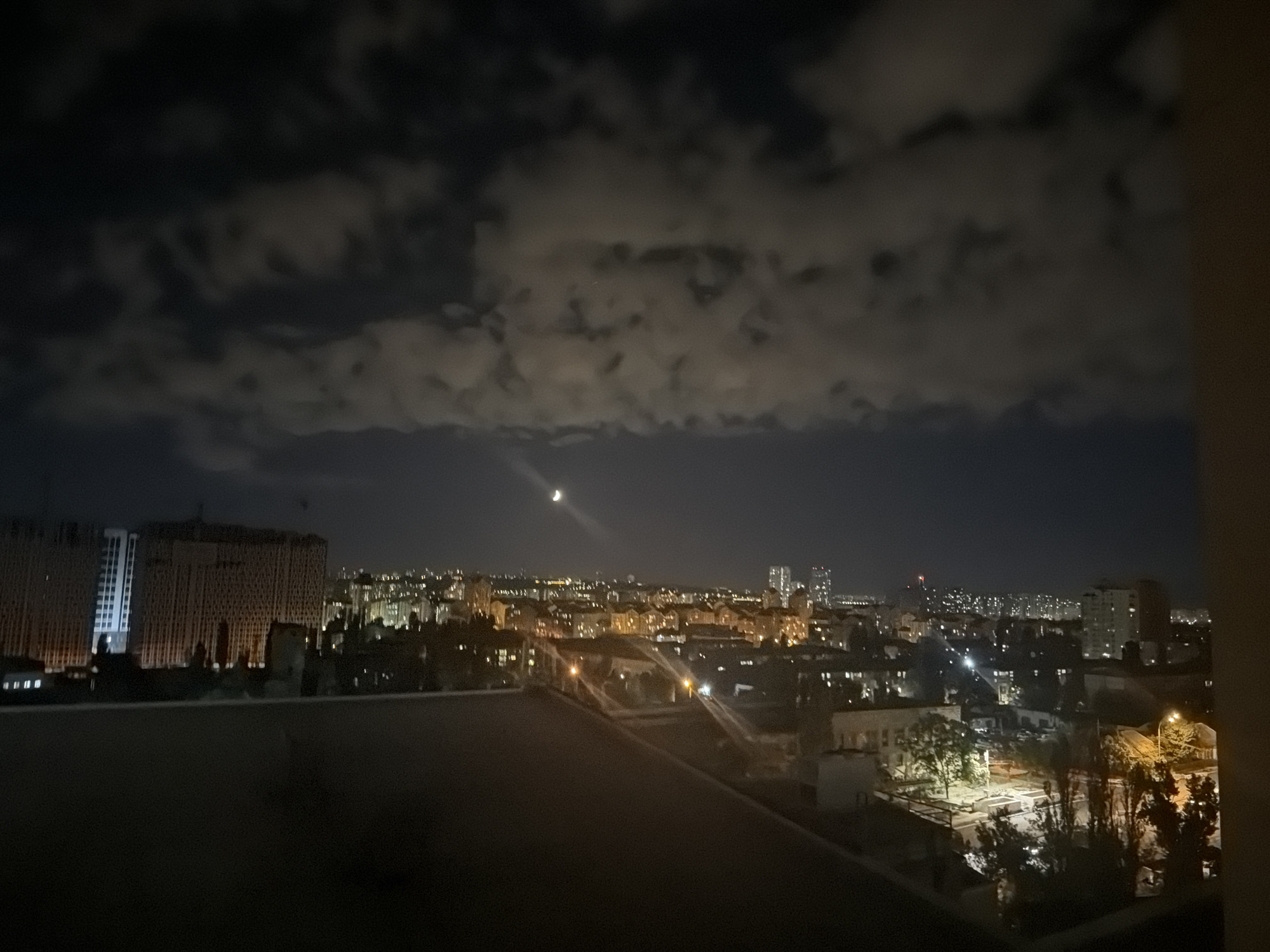 This is a nighttime aerial view of a cityscape, likely in Kyiv, Ukraine, under a cloudy sky with a visible crescent moon. The image captures a dense urban environment characterized by numerous buildings illuminated by artificial lights. In the foreground, a dark, silhouetted structure, possibly a rooftop or balcony, dominates the lower portion of the frame. Beyond this, the mid-ground reveals a sprawling city with varying building heights and architectural styles, many displaying lit windows. A particularly large, modern-looking building with a patterned facade stands out on the left. The background extends to the horizon, showing a more distant cluster of buildings and the soft glow of city lights. The overall atmosphere is dark and subdued, with the moon and city lights providing the primary illumination. There are no people or animals visible in the image.