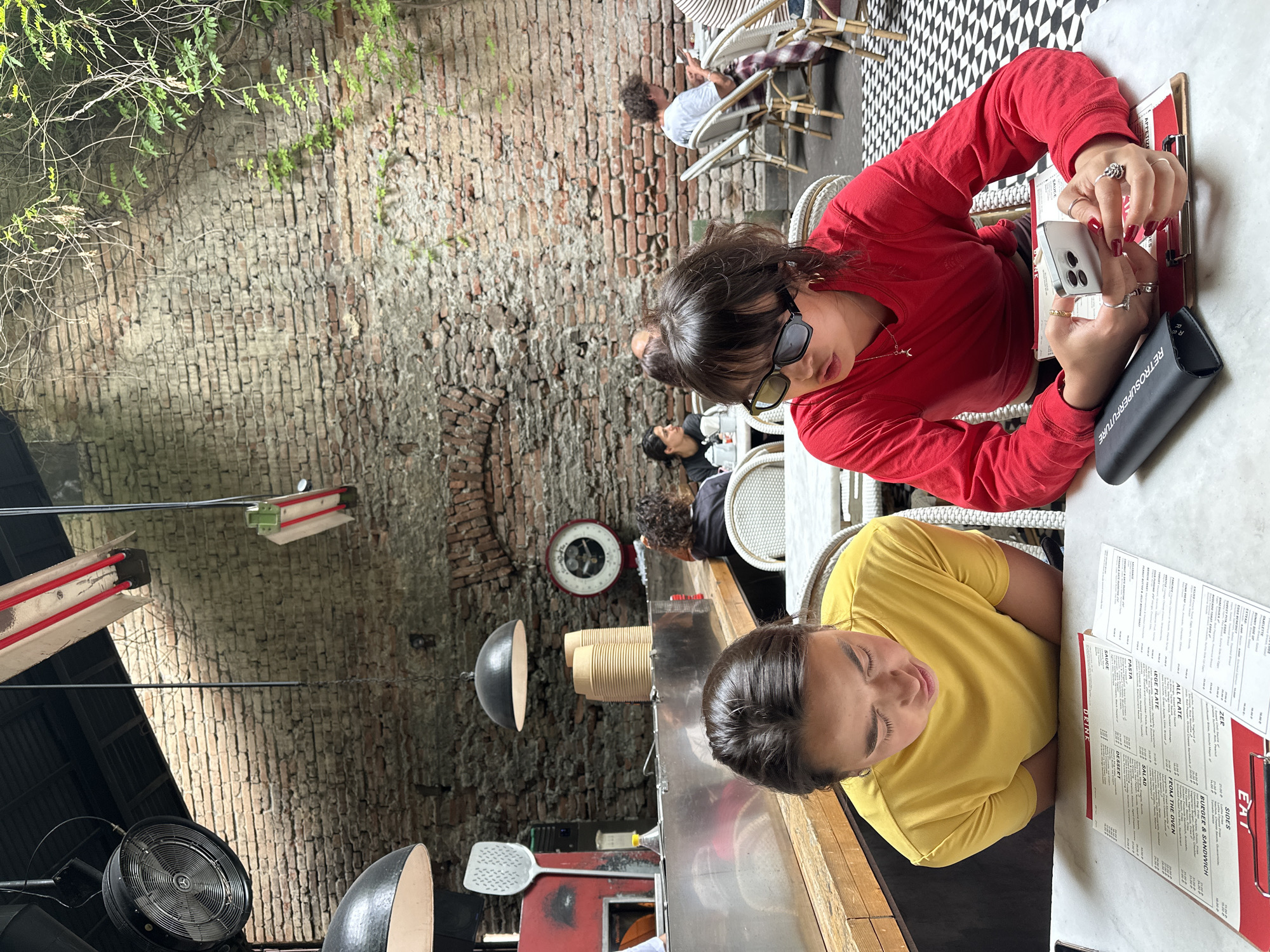 The image depicts an outdoor seating area of a restaurant in Tbilisi, Georgia, characterized by exposed brick walls and an industrial-style ceiling with fans and heating lamps. Two young women are seated at a table in the foreground. The woman on the left, wearing a yellow t-shirt, is looking down at a menu. The woman on the right, wearing a red long-sleeved shirt and black sunglasses, is looking at her phone while holding it in her hands. Various rings are visible on her fingers. A black phone case with the text "RETRO SUPERFUTURE" is on the table next to her. In the background, other patrons are seated at tables, and a food preparation counter is visible with stacks of disposable cups and a pizza peel. The floor has a black and white checkered pattern. The overall ambiance suggests a casual dining experience in a city setting.