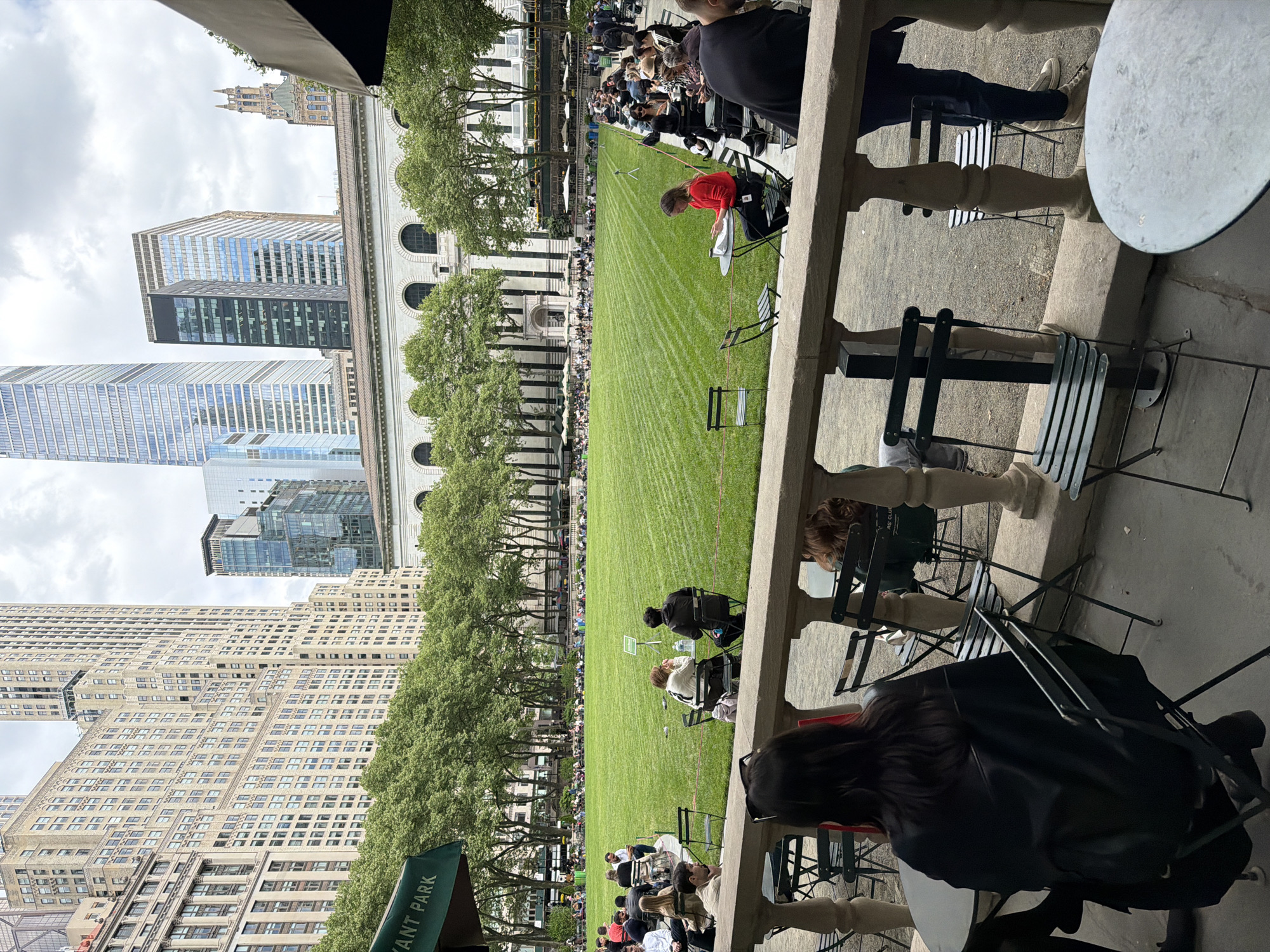 The image captures a view of Bryant Park in New York City. A large, rectangular green lawn stretches through the center of the park, lined with trees on either side. People are seated on chairs spread across the lawn. Tall buildings are visible in the background, including skyscrapers with glass facades and older buildings with architectural details. In the foreground, there are outdoor tables and chairs, with people sitting at some of them. The image is taken from a slightly elevated position, possibly from a building overlooking the park.