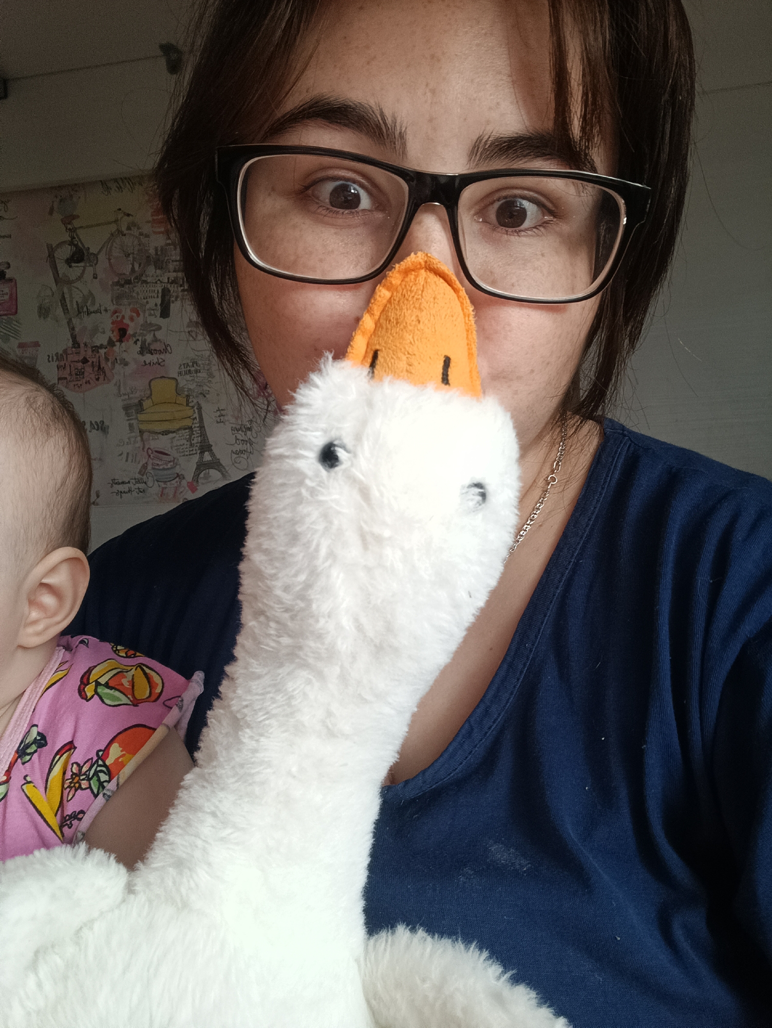 A close-up, informal portrait captures a woman holding a fluffy white toy goose in front of her face. The toy's orange beak covers her nose and mouth, while her wide, brown eyes peer over it through black-rimmed glasses, conveying a playful or surprised expression. She has dark hair, styled with bangs, and wears a dark blue v-neck top with a thin silver chain around her neck.
On the left side of the frame, the top of a baby's head, ear, and shoulder are visible. The baby wears a pink top adorned with a colorful pattern of yellow, orange, and green elements, resembling fruits or flowers.
The background is an indoor wall covered with patterned wallpaper featuring various illustrations and text. Visible elements include "Paris," an Eiffel Tower, bicycles, perfume bottles, and partially legible words such as "ST. A," "NEW YORK," "C'est la vie," "Sweet," and "love." The lighting suggests a daytime setting. The scene depicts a lighthearted, domestic moment in Noviy, Russia.