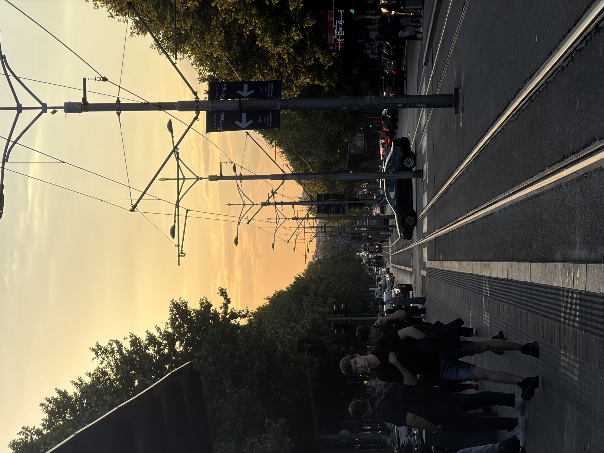 An eye-level, slightly tilted shot shows a street in Vracar Urban Municipality, Serbia, at dusk. The street has a tram line running down the center and is lined with trees on either side. Overhead tram wires are suspended from metal poles along the street. The sky is a gradient of yellow and orange as the sun sets. Cars are parked along the road, and some are driving, creating traffic. On the right side of the frame, a group of young people are walking along the pavement. Street signs are visible on the poles, with the word "Primea" displayed.