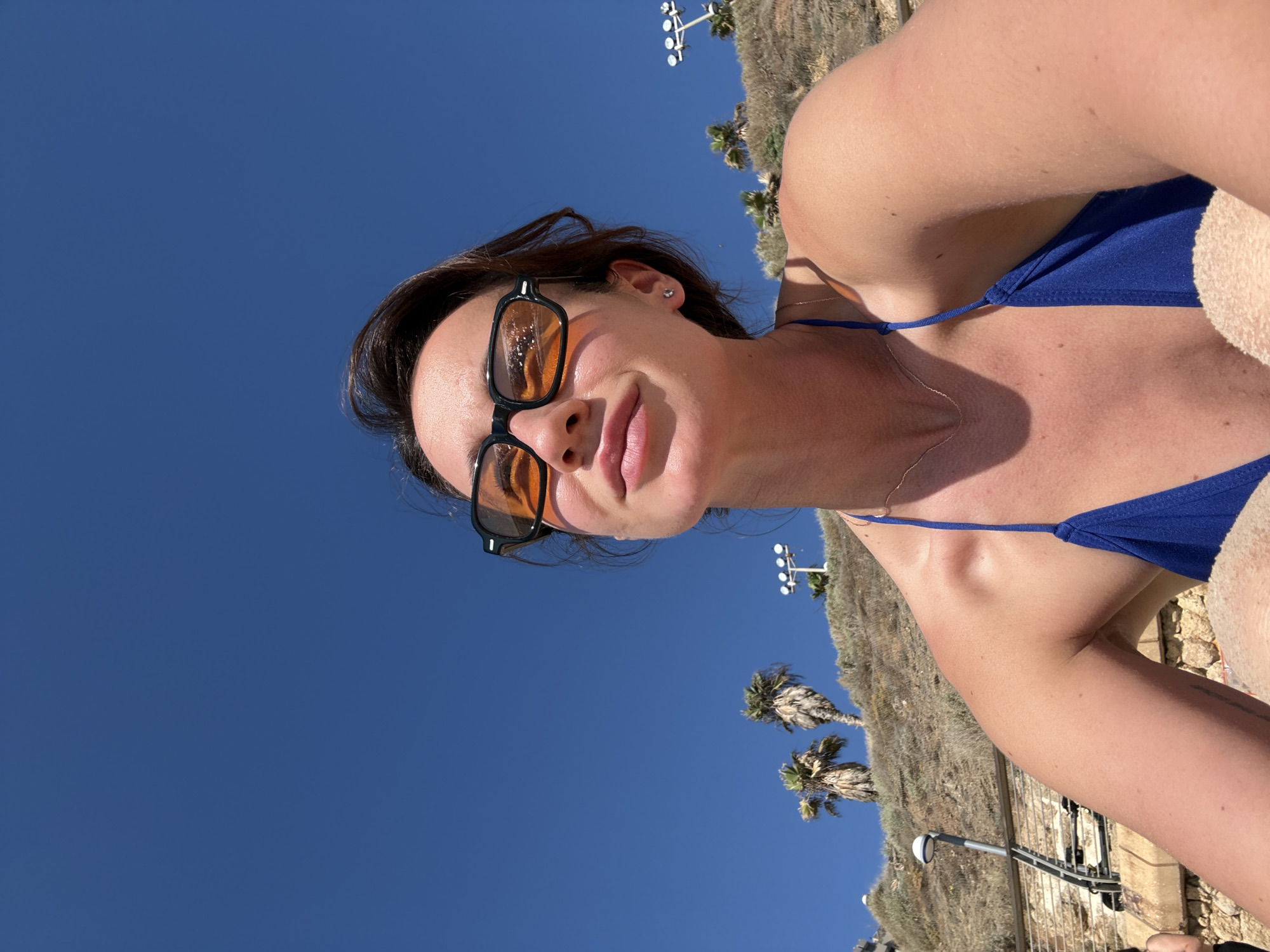 A woman wearing a blue bikini and orange-tinted sunglasses is photographed from a low angle against a clear blue sky. She is looking upwards and to the side, with a slight smile. Her hair is dark brown and pulled back. In the background, there is a sandy embankment with scattered palm trees and what appears to be some kind of lighting fixtures. Part of a metal railing and a stone wall are visible on the left side of the image. The overall scene suggests a sunny day at a beach or a coastal area in Tel Aviv, Israel.
