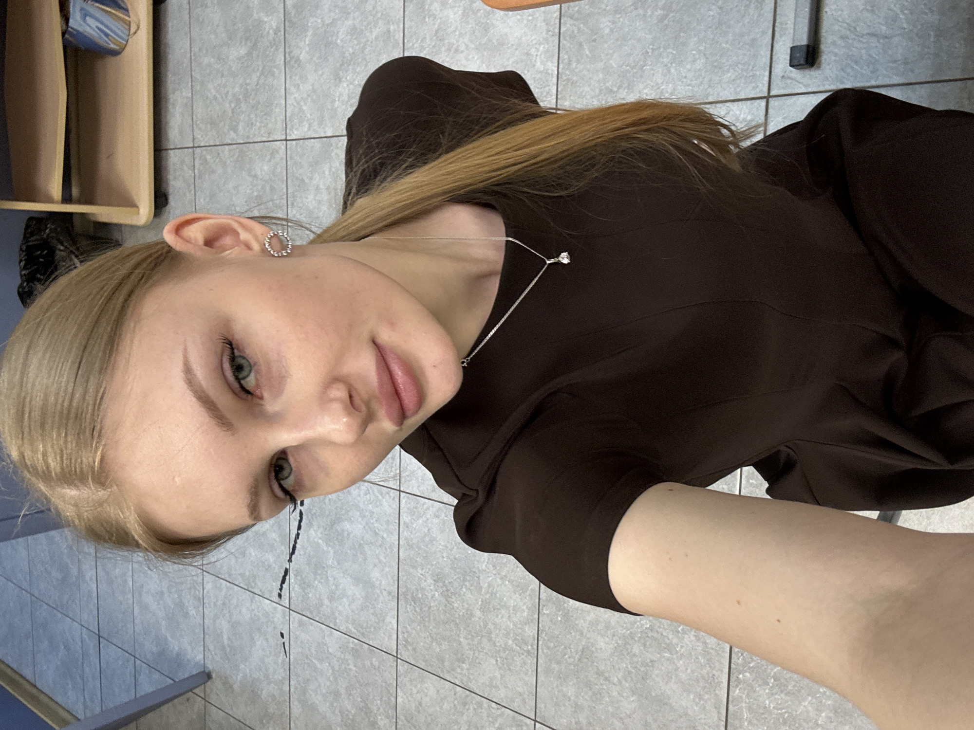 A woman with blonde hair and green eyes, wearing a brown dress and a necklace, is taking a selfie. She has an earring in her left ear. Black liquid is dripping from her left eye, down the tile wall. The background consists of gray tiles and furniture. The location is in Perm, Russia.