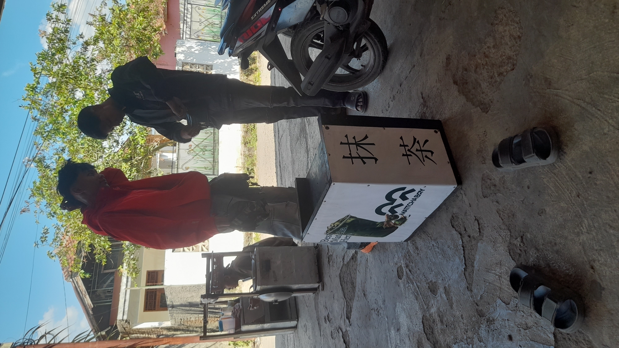 In Selaawi, Indonesia, two young men stand near a matcha vending box on a sunlit, concrete ground. The man on the left wears a red hoodie and grey pants, while the man on the right sports a black jacket and dark pants. In the background, a street vendor's cart and a motorcycle are visible. The scene unfolds on a paved area, possibly an outdoor market or roadside stall, under a bright blue sky dotted with clouds. The environment appears rustic, with weathered concrete and a hint of vegetation.