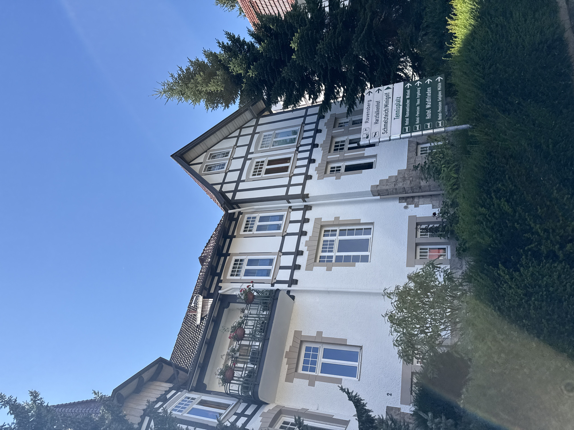 The image shows a building in Bad Sachsa, Germany. The building is white with dark timber framing. There is a balcony with plants on it and several windows visible. In front of the building is a signpost with multiple directional signs. The signs indicate directions to Ravensberg, Harzfalkenhof, Schmelzteich/Minigolf, Tennisplatz, Hotel Romantischer Winkel, Hotel-Pension Haus Irgebrug, Hotel Waldfrieden, and Hotel-Pension Gelpkes Mühle. There are also trees and bushes in front of the building. The sky is blue and clear.