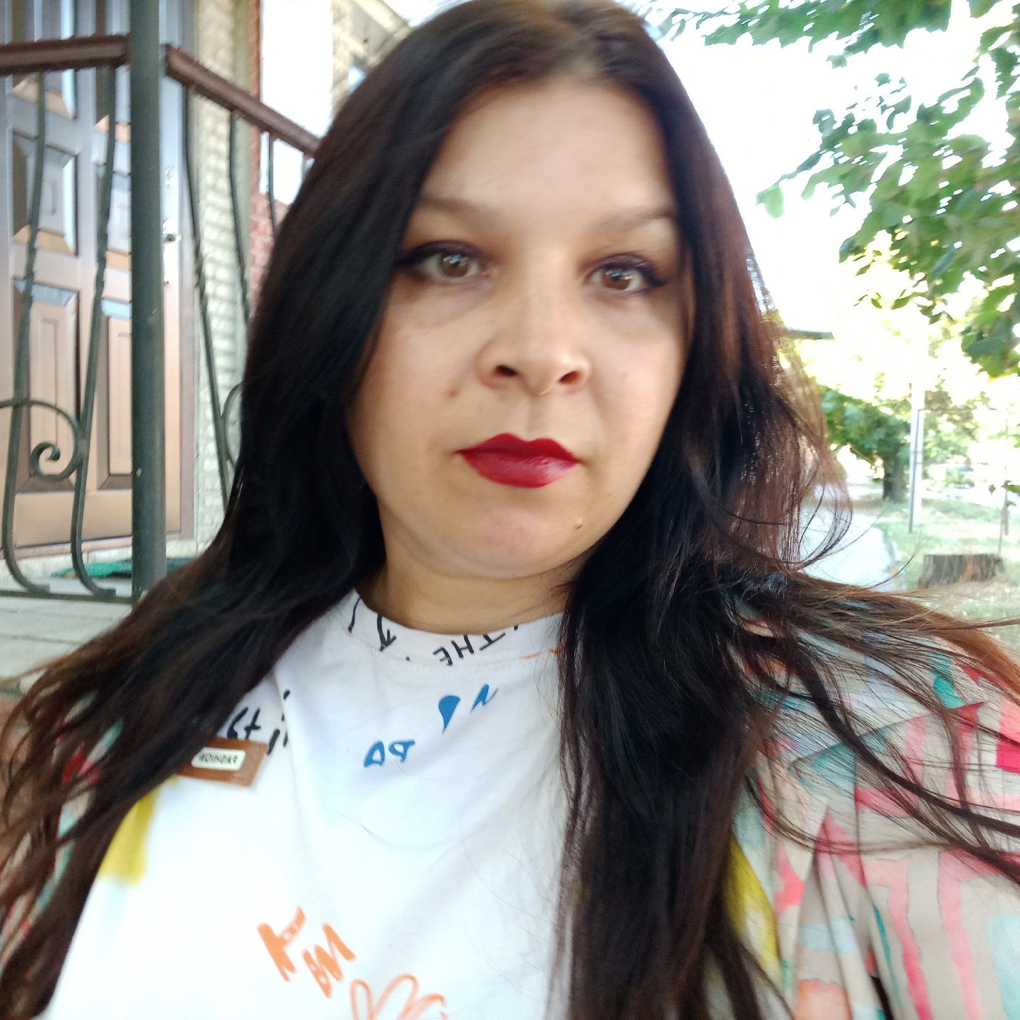 A medium shot shows a woman with long, dark hair and red lipstick looking at the camera. She is wearing a white t-shirt with abstract colorful designs and a colorful patterned jacket. A dark metal railing and a doorway are visible behind her. The background also includes trees and foliage, suggesting an outdoor setting, possibly in a park or yard. There is a small brown tag with text on her t-shirt. The lighting suggests it is daytime.