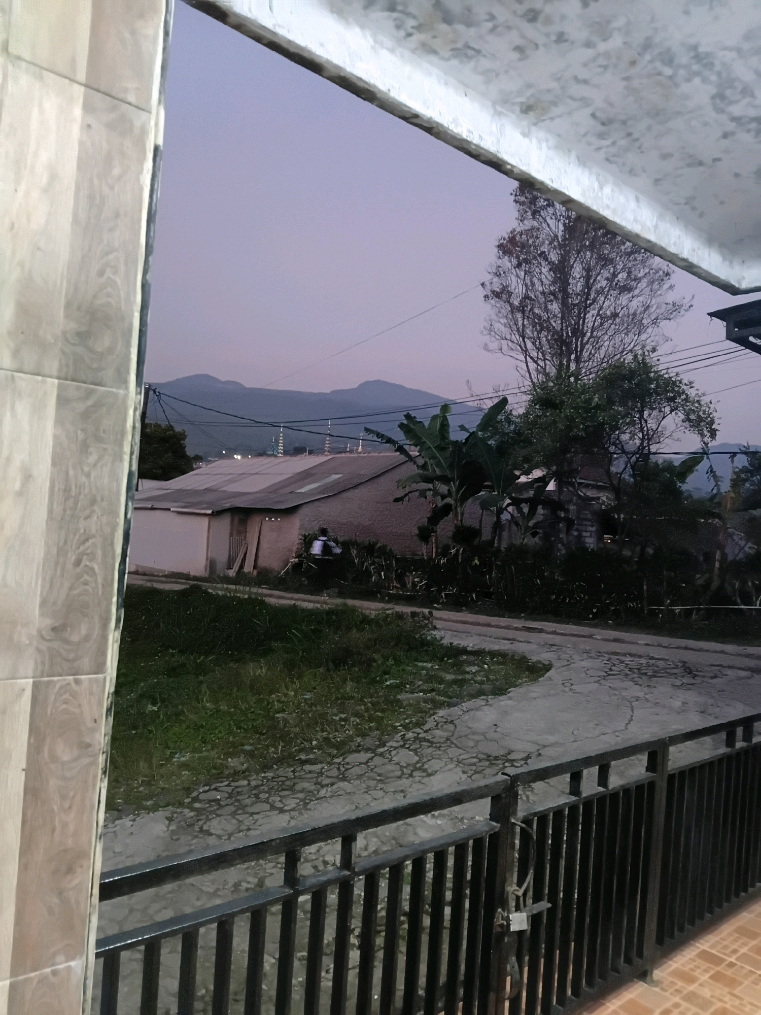 The image depicts a serene twilight scene in Ciater, Indonesia, viewed from a covered veranda. In the foreground, a black metal railing with a padlock secures the edge of the veranda, while the weathered, light-colored tiles of the floor are visible. Beyond the railing, a patch of lush green grass transitions into a winding concrete pathway.
In the middle ground, a low-slung building with a corrugated metal roof is situated, accompanied by a tall, barren tree with visible branches. Banana trees and dense foliage add a tropical element to the landscape. A single individual, wearing a white shirt, is visible near the building, seemingly engaged in an activity. Power lines crisscross the sky, hinting at the presence of infrastructure.
In the background, a chain of mountains stretches across the horizon under a soft purple sky, suggesting a rural or semi-rural setting. Several minarets, characteristic of mosques, pierce the skyline, indicating a community with religious structures. The overall impression is one of peaceful, rural evening in Ciater.
