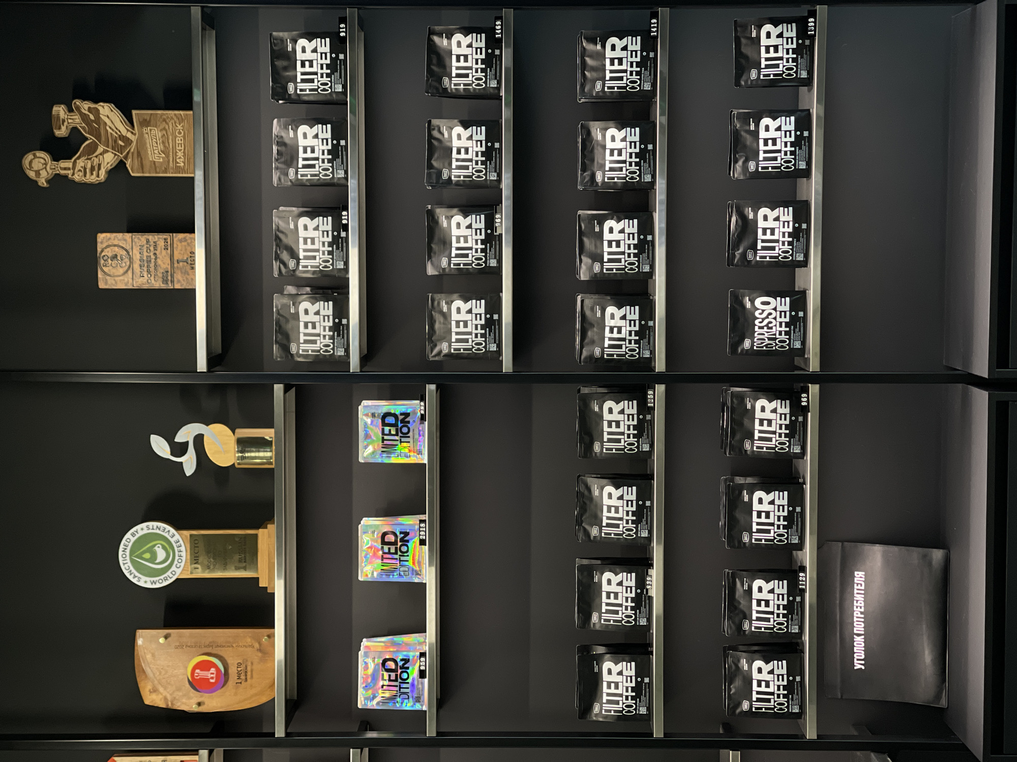 The image shows a retail display of coffee products and awards, likely in a specialty coffee shop. The display is organized on dark gray shelves against a black wall.
On the top shelf, there are several awards and trophies. One is a wooden plaque with a circular logo and text, appearing to be a first-place award. Next to it is a larger trophy with a circular emblem that reads "SANCTIONED BY WORLD COFFEE EVENTS" and "1 МЕСТО". To the right of that is another award, possibly a stylized coffee plant. Further to the right, on the second level of awards, is a wooden plaque depicting a hand holding coffee brewing equipment and text that appears to include "КОФЕЙНЫЙ ПАТРУЛЬ" and "ИЖЕВСК", indicating a connection to Izhevsk, Russia.
Below the awards, the shelves are filled with bags of coffee. On the third shelf from the top, there are three bags labeled "LIMITED EDITION" with holographic packaging. Below these, on the fourth, fifth, and sixth shelves, are numerous black bags of coffee labeled "FILTER COFFEE". Some of these bags have small labels indicating prices. On the sixth shelf, there is one bag labeled "ESPRESSO COFFEE".
On the bottom shelf, there is a dark gray folder or sign with white Cyrillic text that reads "УГОЛОК ПОТРЕБИТЕЛЯ," which translates to "Consumer Corner."
The overall impression is a well-organized and modern retail environment showcasing high-quality coffee products and recognition for achievements in the coffee industry, with a specific mention of Izhevsk, Russia.
