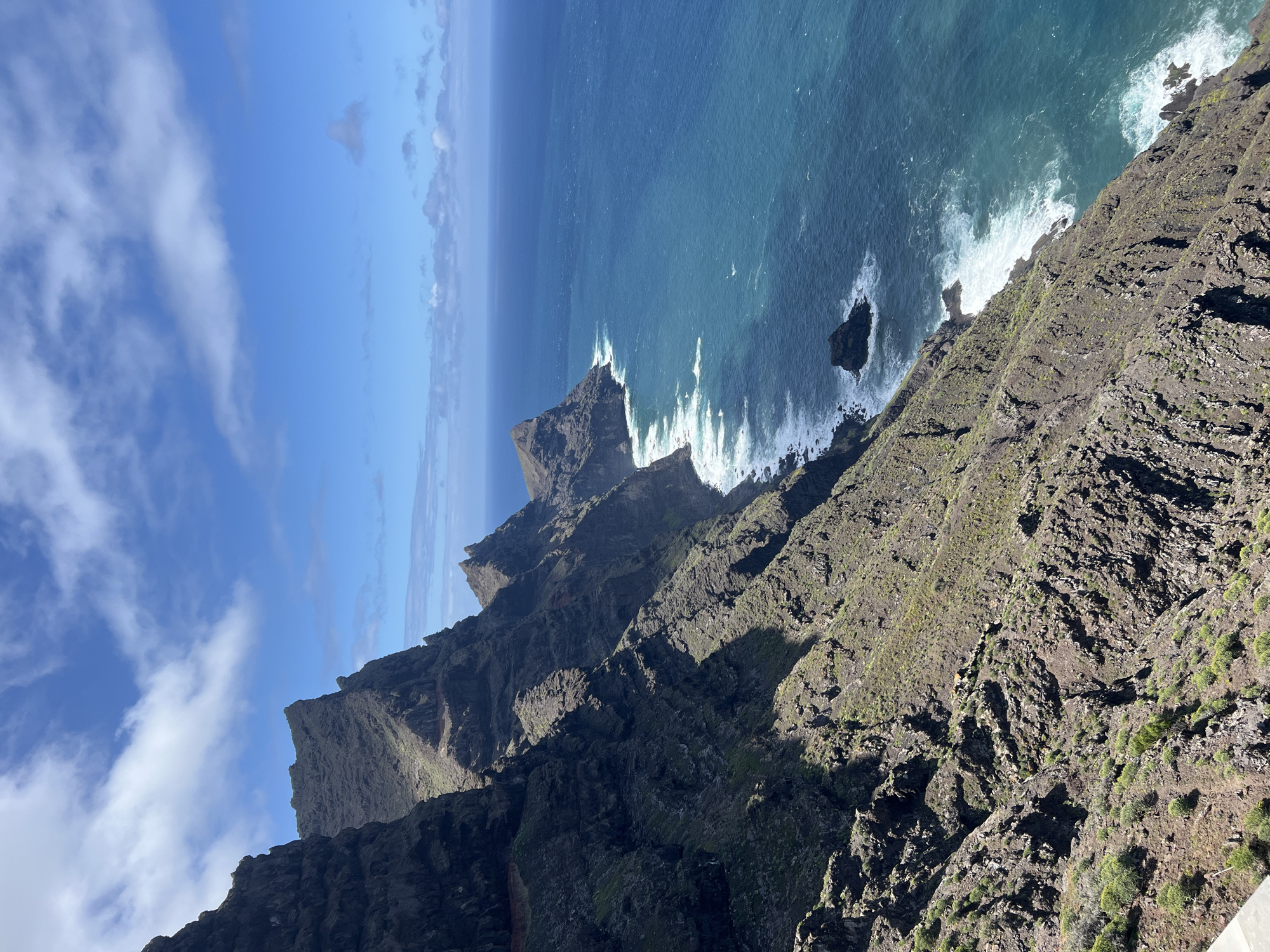 The image shows a steep, rocky coastline meeting the ocean under a partly cloudy sky. Jagged cliffs descend sharply into the water, where waves crash against the rocks. The cliffs are covered with sparse vegetation. The location is Tirma, Spain.