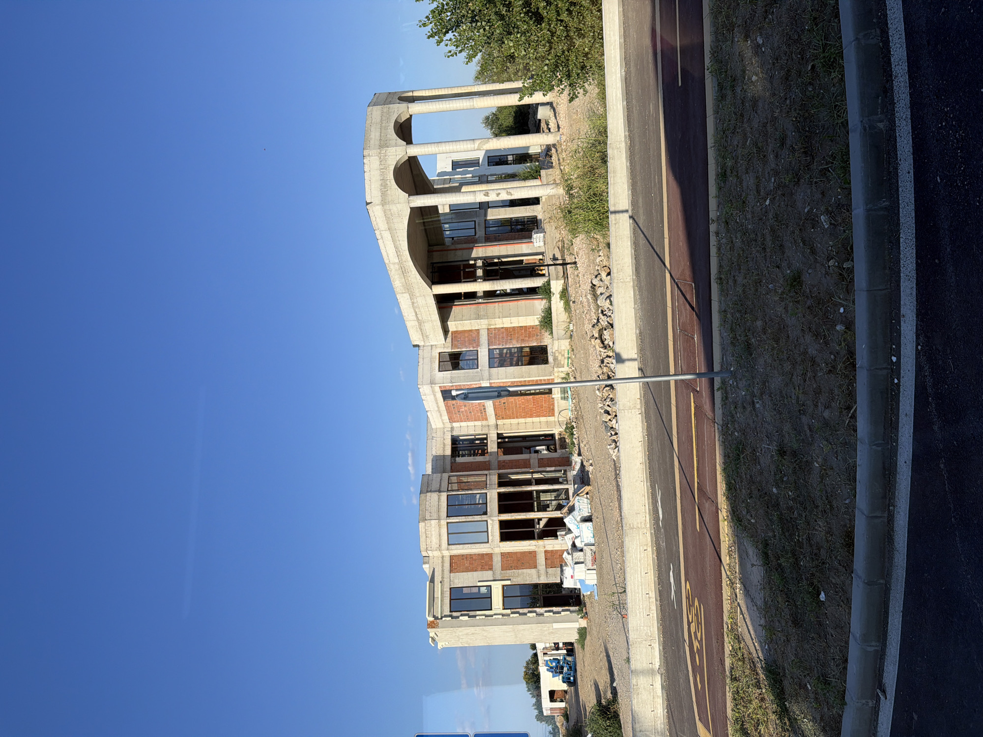 The image shows an unfinished building with a modern architectural design, featuring large windows and exposed brickwork. The building is surrounded by construction materials, including stacks of white insulation panels and piles of rubble. A blue construction lift is visible in the background. The scene is set against a clear blue sky and a landscape with dry vegetation and a road with designated bike and pedestrian lanes. The image appears to be taken from a moving vehicle, as indicated by the slight motion blur and the perspective. The style of the architecture and the urban development suggest a location within a modern residential or commercial area. However, without additional information, it is not possible to determine the specific location within the city of Plovdiv, Bulgaria.