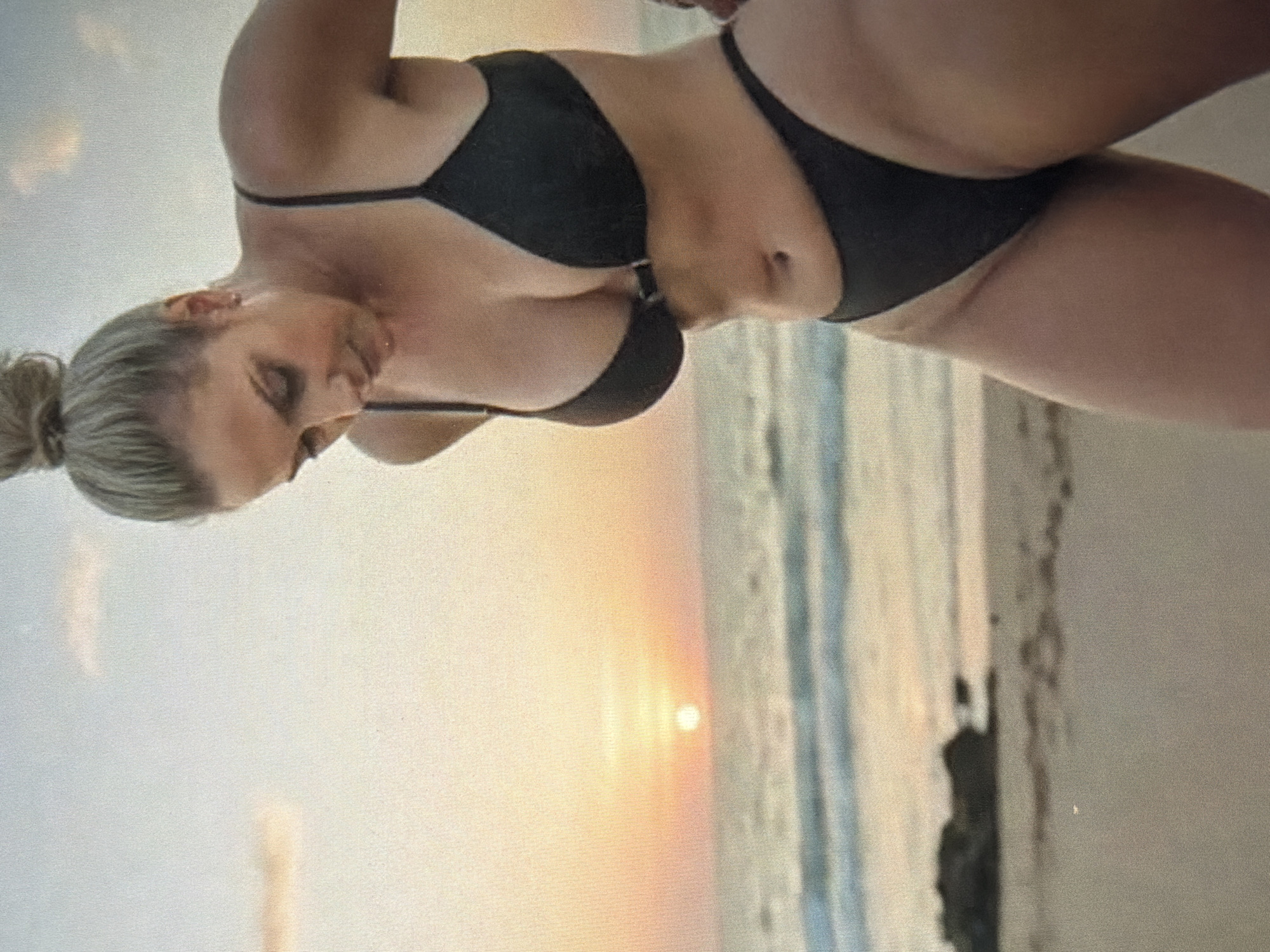 A woman wearing a black bikini stands on a sandy beach at sunset. The sun is low in the sky, casting a warm glow on the scene. The ocean is calm with gentle waves lapping at the shore. In the background, there are some rocks and sand dunes. The woman is looking down, and her hair is pulled up in a bun. The image is taken from a medium shot, focusing on the woman and the beach.