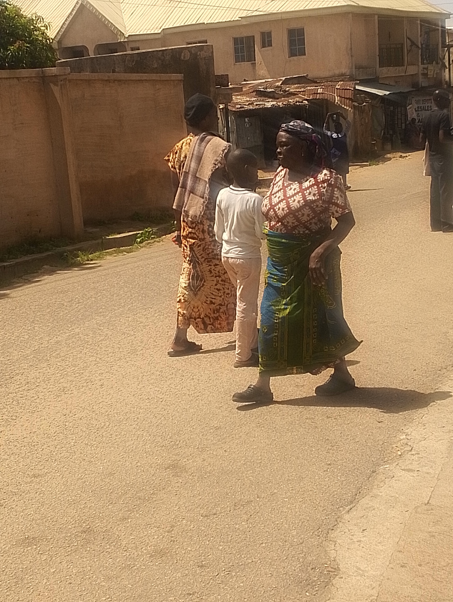 The image depicts a street scene in Jos, Nigeria. Three people are visible on an asphalt road. In the foreground, a person wearing a white long-sleeved shirt and light-colored pants is walking away from the camera. To their right and slightly behind, an adult is wearing a red and white patterned shirt and a blue and green patterned wrap skirt. This person is facing left and has their right hand on their hip. To the left and behind the person in white, another adult is wearing a tan and brown patterned garment and a dark head covering. This person is facing away from the camera. In the background, buildings with corrugated metal roofs and other structures are visible. A sign that says "LE SALES & AL" is partially visible on one of the buildings. The overall environment suggests a sunny day in an urban area.