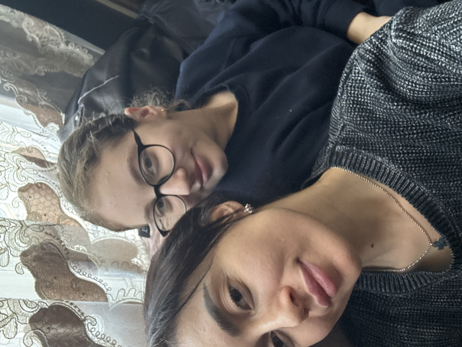 This image depicts two females in a close-up, indoor setting within the city of Bolnisi, Georgia. The subject in the foreground, an adult female with dark hair, wears a dark grey knitted sweater and a silver-toned chain with a small, dark, possibly cross-shaped pendant. A silver-toned earring is visible in her left ear. She looks directly at the camera with a slight smile. Behind and to the right, a younger female with lighter, curly hair wears black-framed glasses and a dark blue long-sleeved top. A small gold-toned hoop earring is visible in her left ear. She also looks towards the camera with a faint smile. The background features a light-colored curtain with a brown, ornate pattern and a dark, indistinct object.