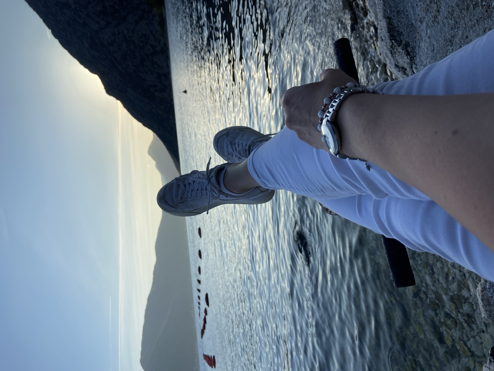 A person is sitting on what appears to be the edge of a structure or embankment, with their legs dangling over the water. They are wearing white pants and light gray sneakers. The person has a watch and a bracelet on their left wrist, and they are holding a black cylindrical object in their left hand. The water has ripples, suggesting a gentle breeze or current. In the background, there is a mountain range, partially obscured by haze or fog. The sky is light blue, transitioning to a lighter shade near the horizon, possibly indicating sunrise or sunset.
It is not possible to confirm if the location is in the city of Donji Orahovac, Montenegro from the provided image alone.