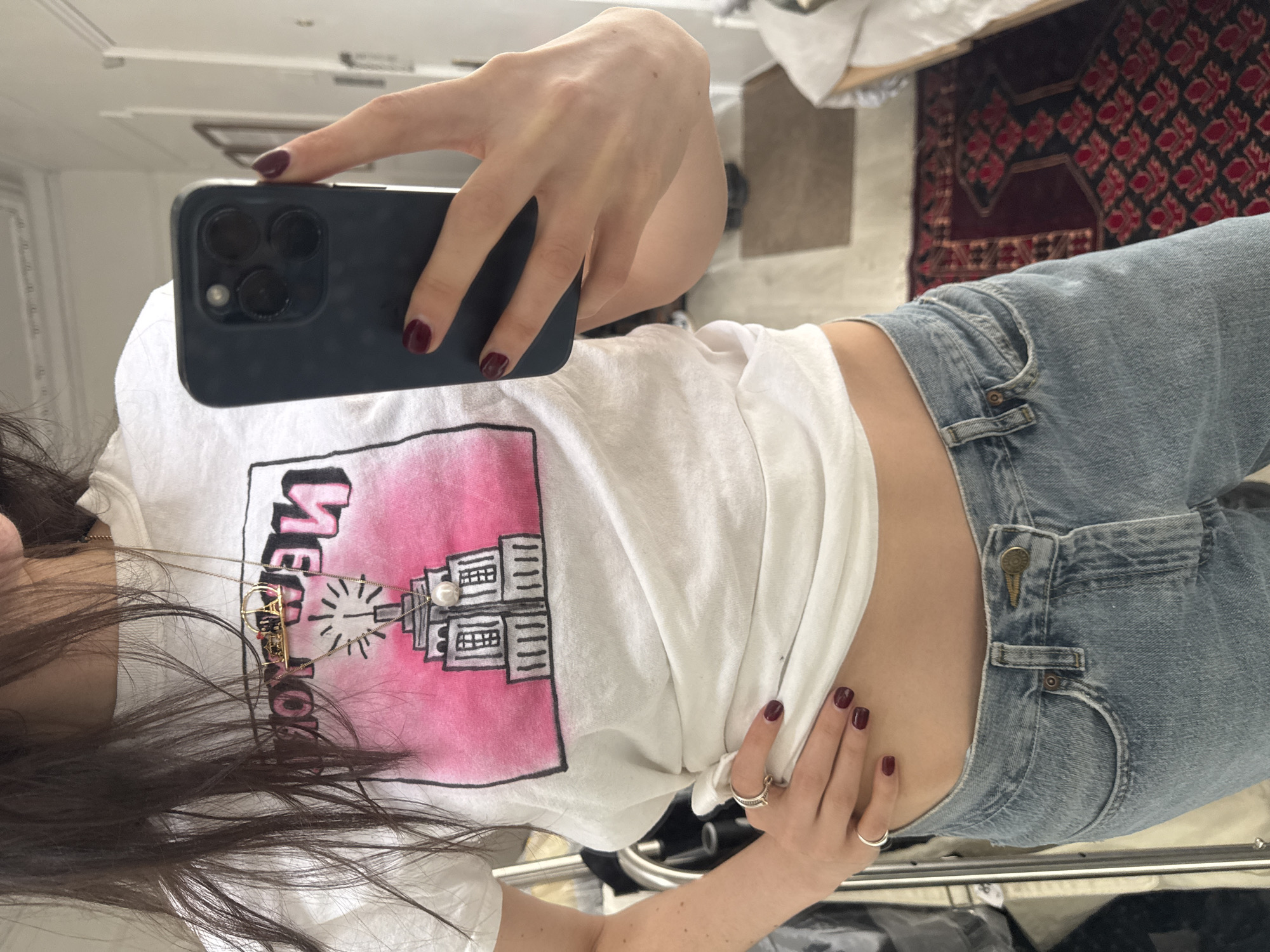 A woman in blue jeans and a white t-shirt with a drawing of the Empire State Building takes a selfie with a black phone. She wears dark-red nail polish and two silver rings. The background includes a patterned rug, a chair, and a white wall. The location is inferred from the t-shirt to be the city of New York, United States of America.