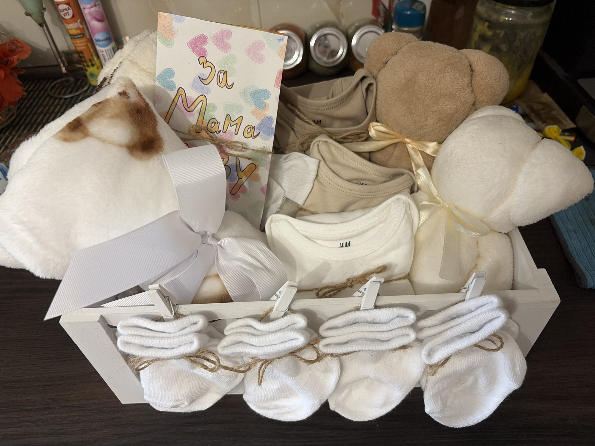 This is a medium shot, eye-level, indoor photograph of a gift basket filled with baby items and arranged on a dark wooden surface. The gift basket is made of white wood and contains several baby clothing items, including onesies in beige and white, and four pairs of white baby socks folded and tied with twine. A white ribbon is tied in a bow on the left side of the basket, and a card with colorful hearts and text in Cyrillic script is visible. There are also two plush toys, a beige teddy bear and a white plush elephant or similar animal, positioned in the basket. In the background, on a shelf, are various items including jars, tubes of what appear to be vitamins or supplements, and a blender. The lighting is moderate and appears to be artificial. The scene does not provide any specific visual cues to determine its location within the city of Burgas, Bulgaria.