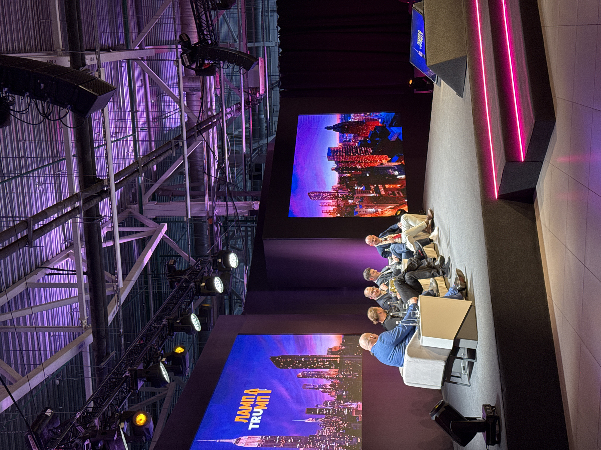 The image shows a conference or summit taking place indoors. Six people are seated on a stage behind a low table with water bottles. Two large screens flank the group displaying images of a city skyline, possibly New York, overlaid with text that appears to be "ЛАМПА TRUMI." A complex network of metal beams, pipes, and lighting fixtures is visible on the ceiling above the stage. A set of steps with pink neon lights adorns the right side of the stage. The location is likely Perm, Russia, as indicated by the text on the screen and the overall setting of the event.
