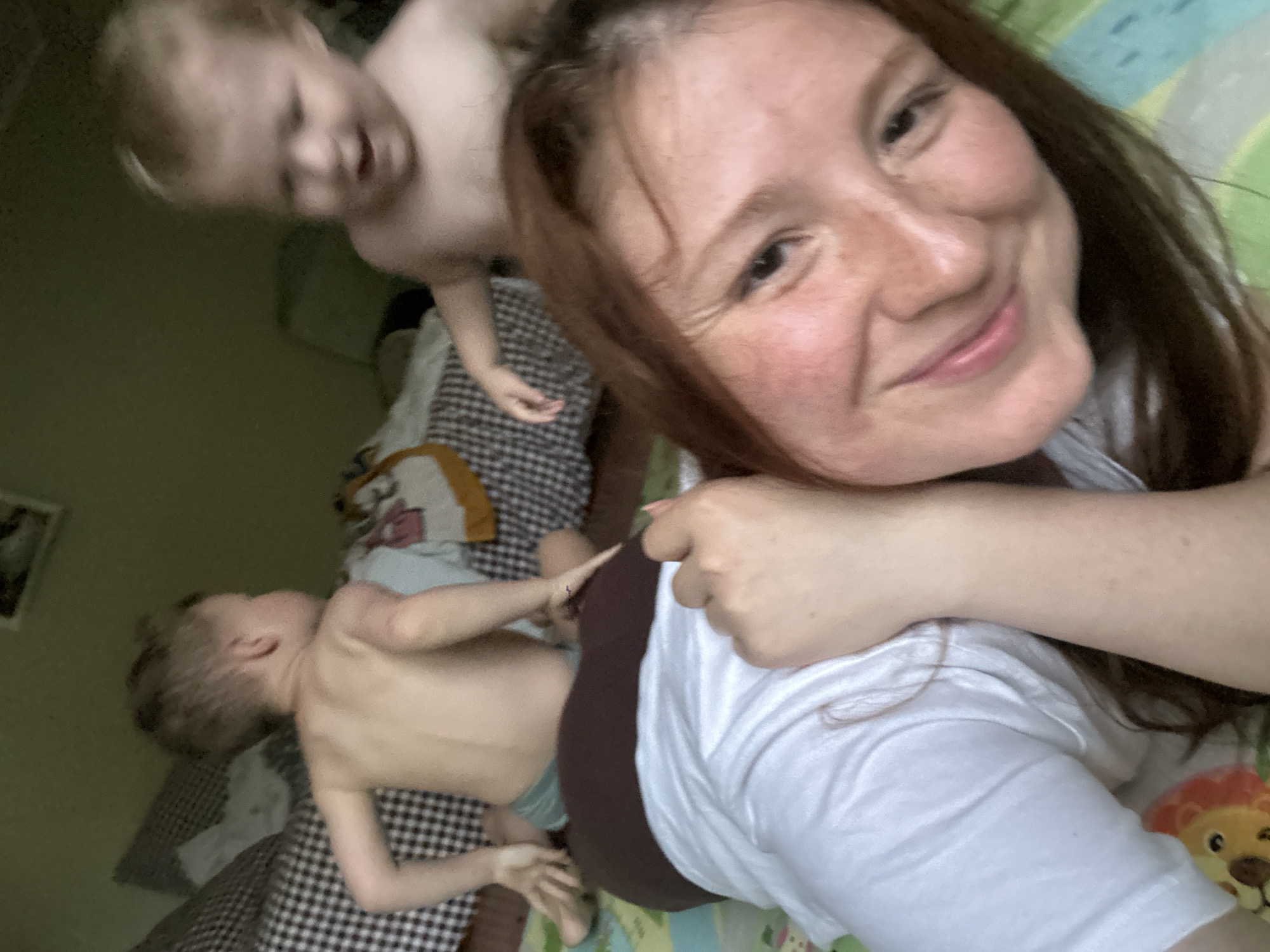 A woman is taking a selfie with two children, one lying on a bed and the other standing over them. The location may be a private home in Nizhny Novgorod, Russia, but lacks definitive regional identifiers for confirmation. The woman has reddish-brown hair and freckles. She is wearing a white shirt. The child on the bed is wearing underwear. The bed has a checkered blanket. A stuffed lion is visible on the bed.